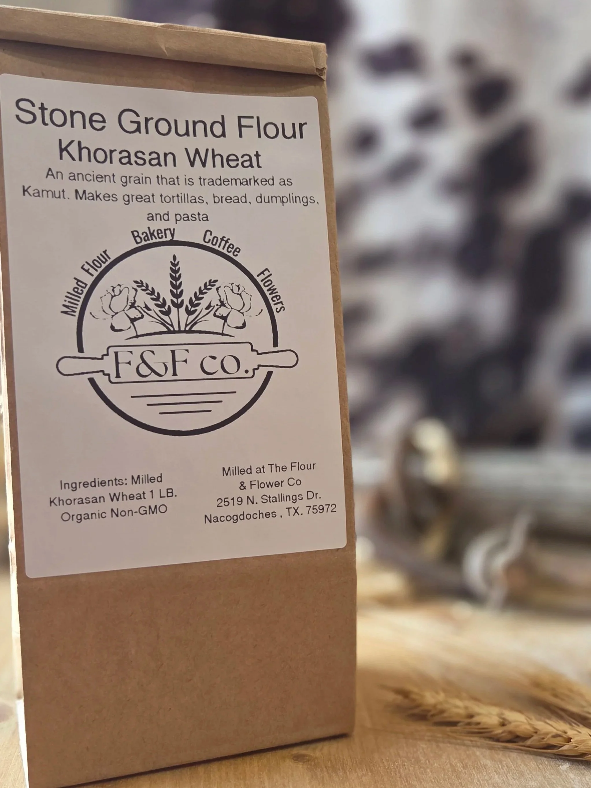 A brown paper bag with a white label featuring black text that reads "Stone Ground Flour Khorasan Wheat" and a circular logo with flowers and wheat, and the words "Milled Flour," "Bakery," "Coffee," and "Flowers." The label also provides ingredient and milling information, including the address in Nacogdoches, Texas.