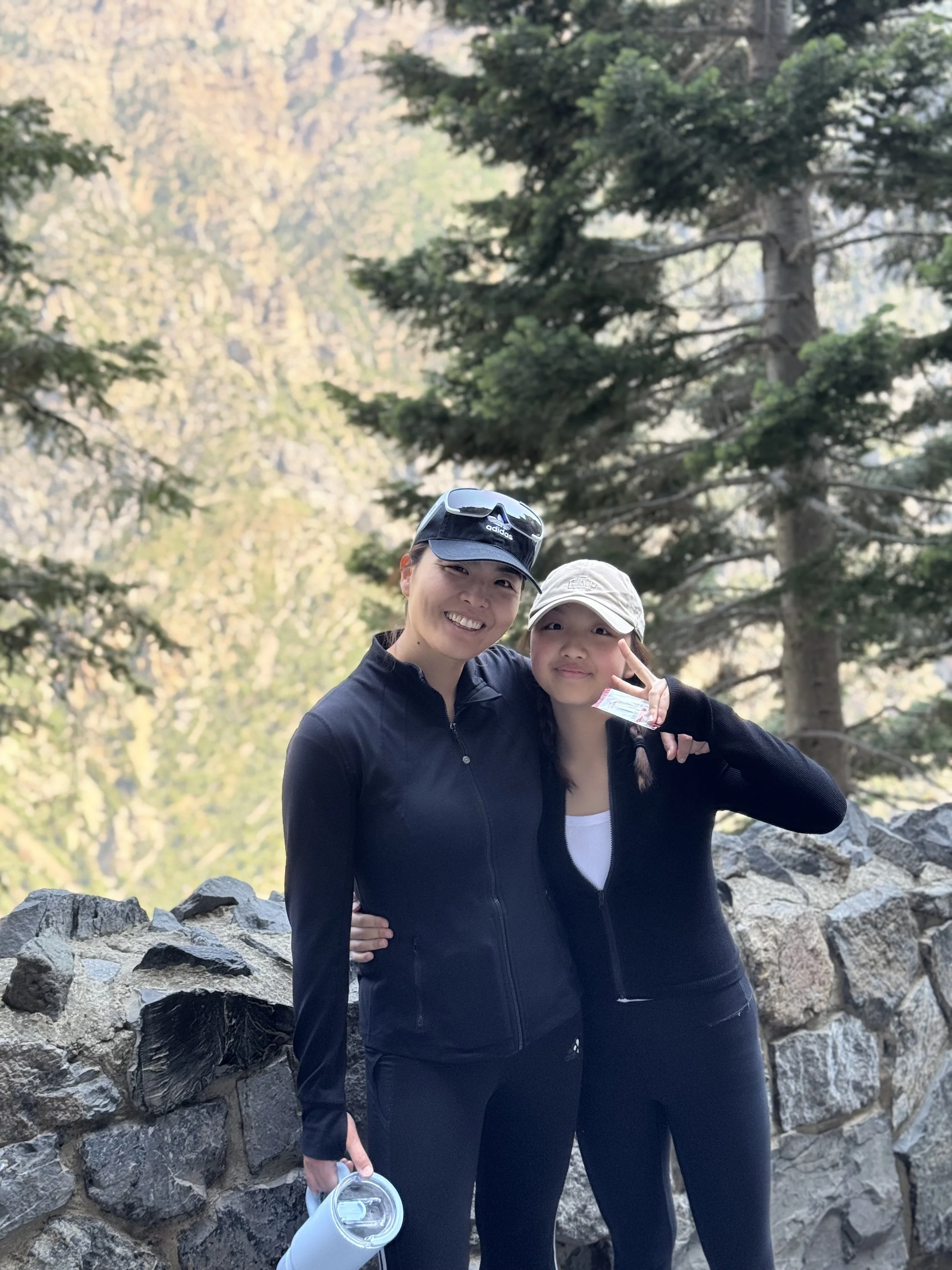 Linda in athletic clothing standing together outdoors, smiling, with trees in the background. One woman is wearing a black cap and sunglasses on her head, and the other is making a peace sign with her fingers. They are standing next to a stone wall.