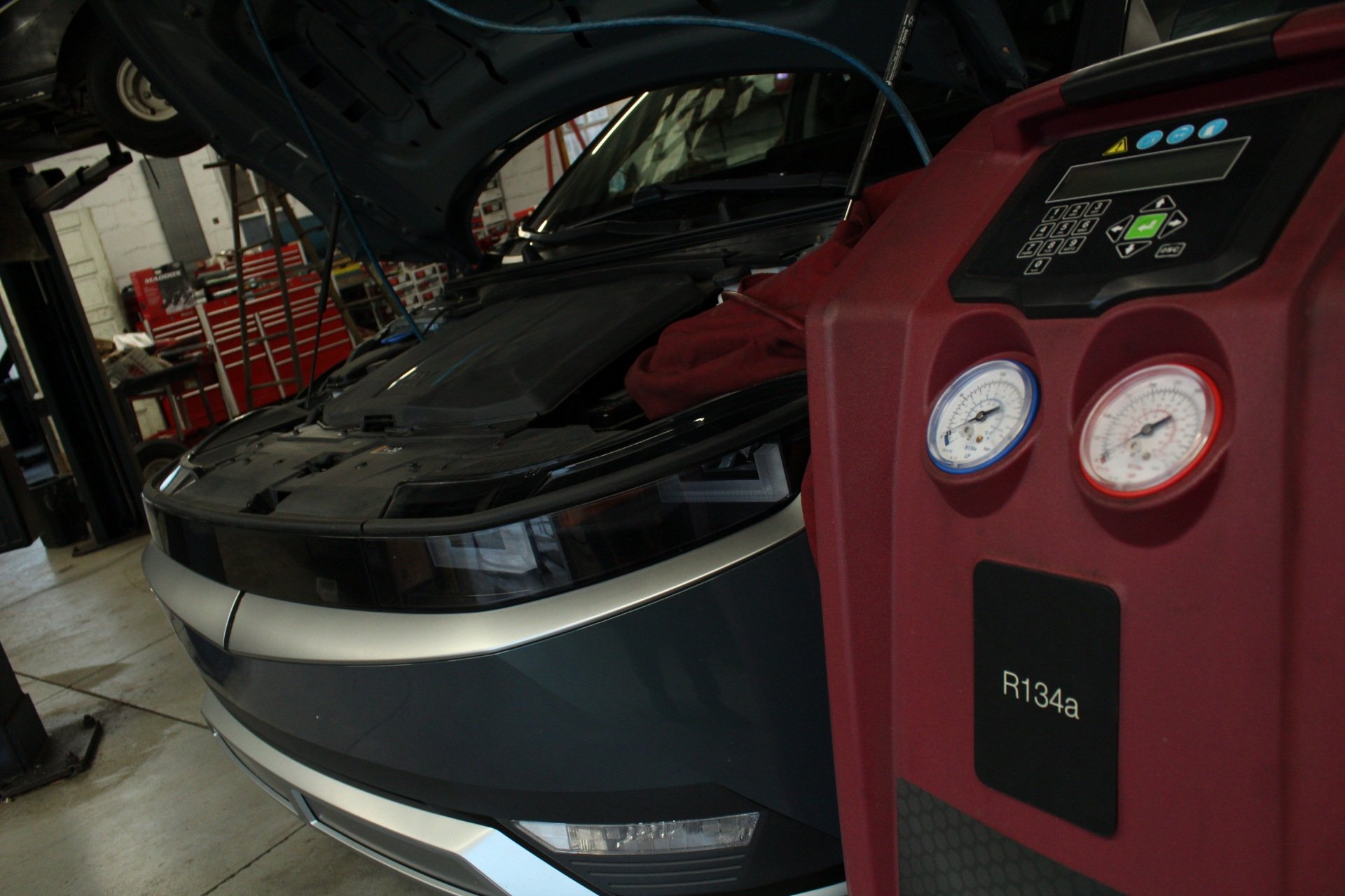 A red car undergoing air conditioning service with hoses connected, and a yellow AC service machine with pressure gauges and controls in a garage.
