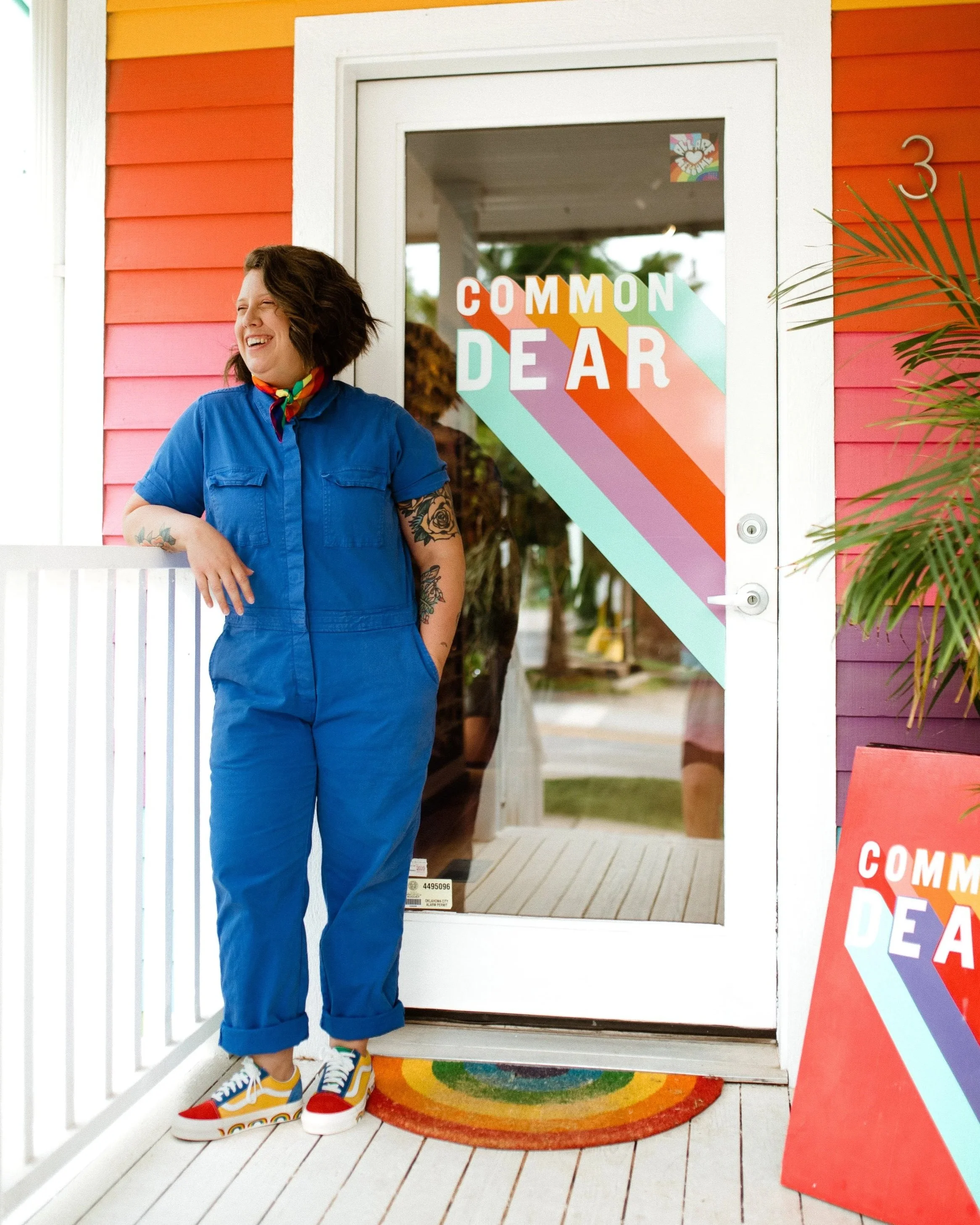 Kelli Crockett Newsome with short dark hair, tattoos, and a colorful scarf around her neck, standing outside a decorated house with a sign that reads 'COMMON DEAR', wearing a blue jumpsuit and colorful sneakers, smiling and looking to her left.