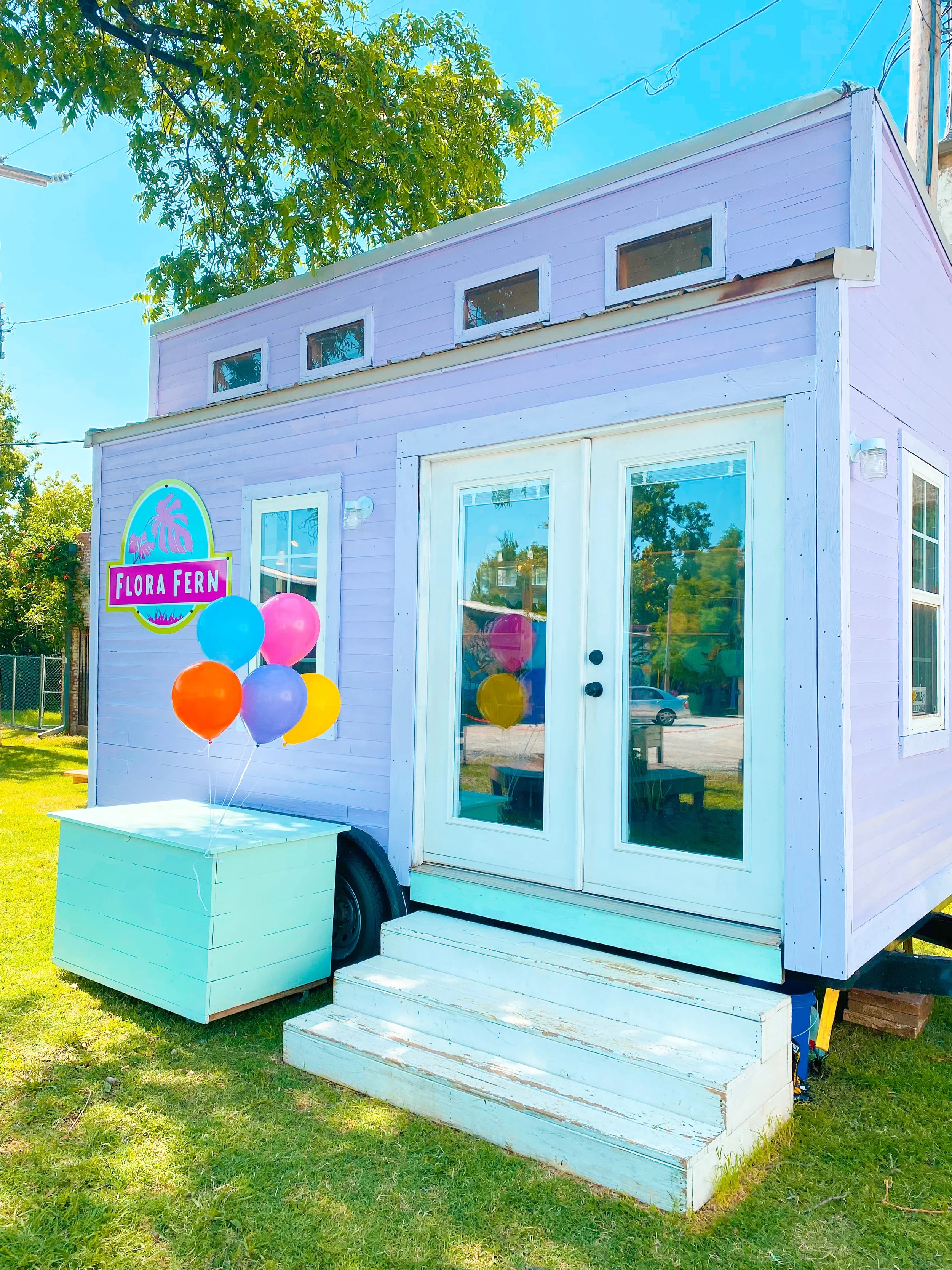 A small, adorable, lavender-colored building with large glass doors, a step, and a sign that reads 'Flora Fern' with a pink background and a purple palm tree logo. The building has a small collection of colorful balloons (blue, pink, purple, yellow, orange) tied to a white rectangular container outside, and is set in a grassy area with trees and a street reflected in the glass.