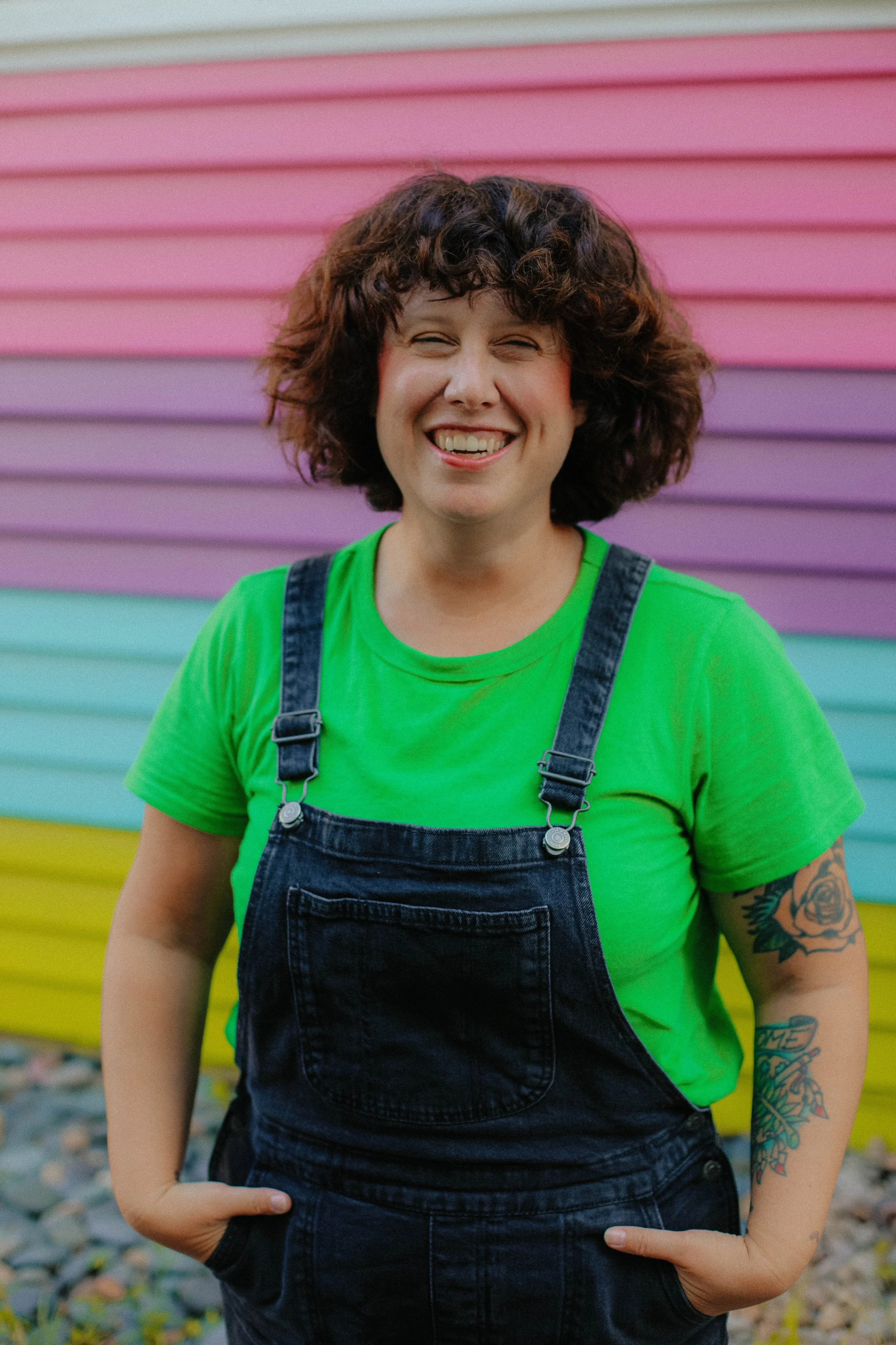 A woman with curly hair, wearing a bright green shirt and denim overalls, smiling in front of a pink and purple rainbow-colored wall.