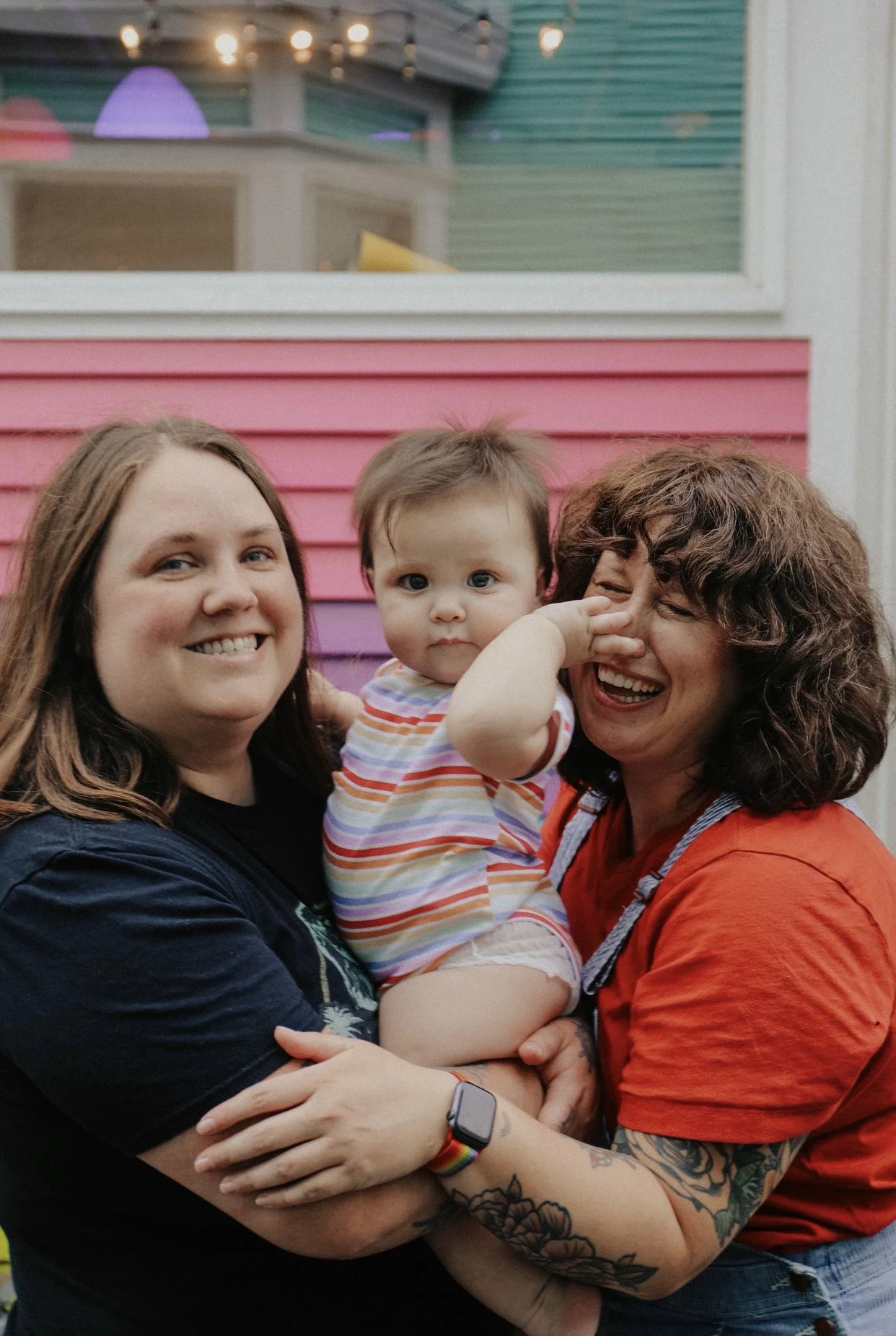 Three people, two women and a young child, smiling and posing together outside near a pink and purple building.