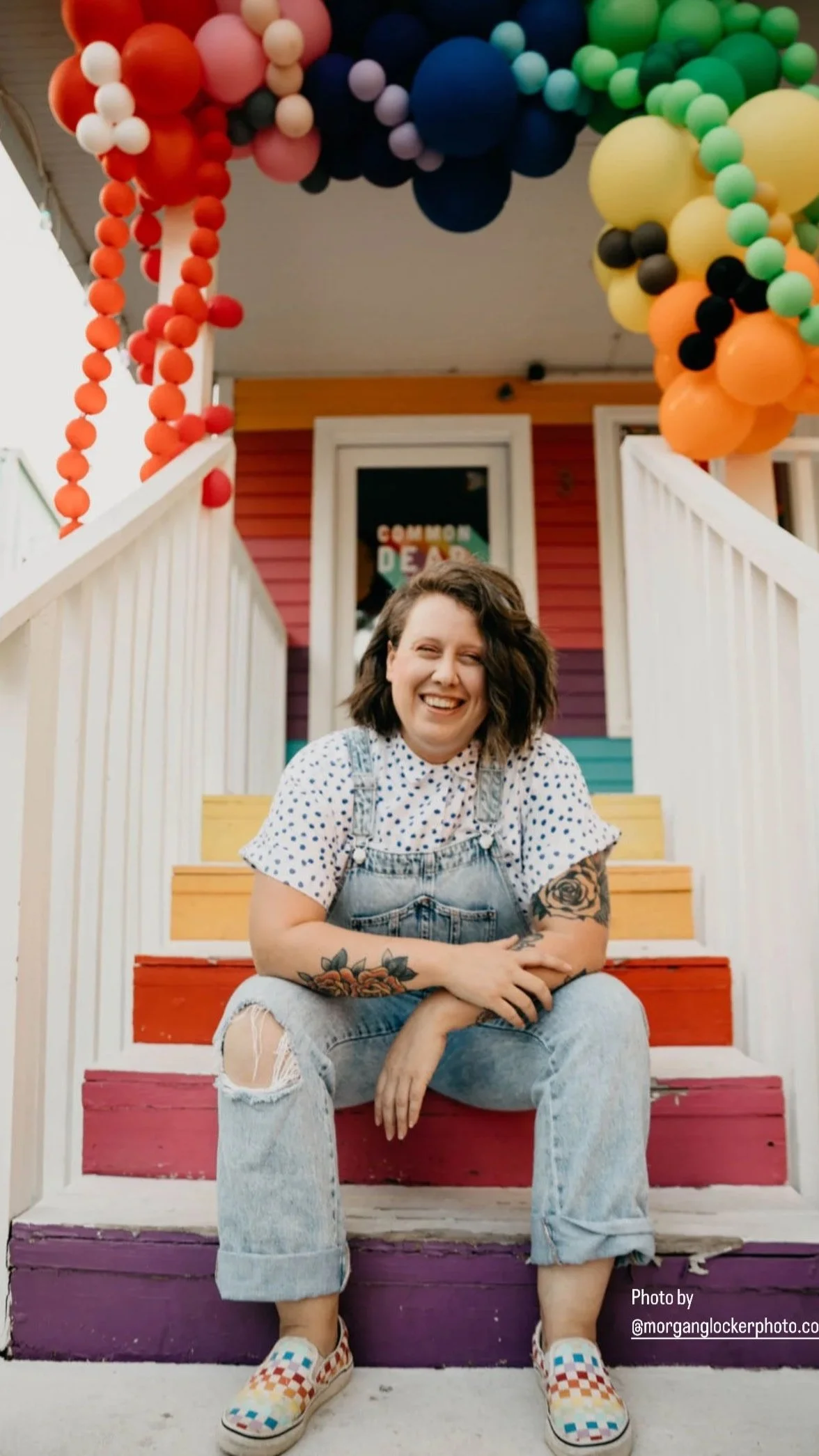 A woman with tattoos sitting on colorful painted steps in front of a decorated house with balloons and a sign that says 'Cannon Deda.' She is smiling and wearing a white polka dot shirt, denim overalls, and checkered slip-on shoes.
