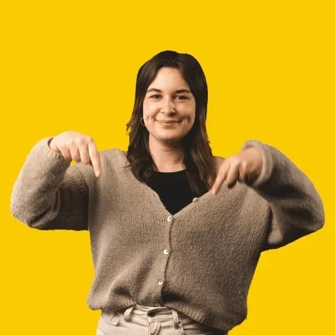 A woman with dark hair smiling, pointing at her oversized beige cardigan against a solid yellow background.