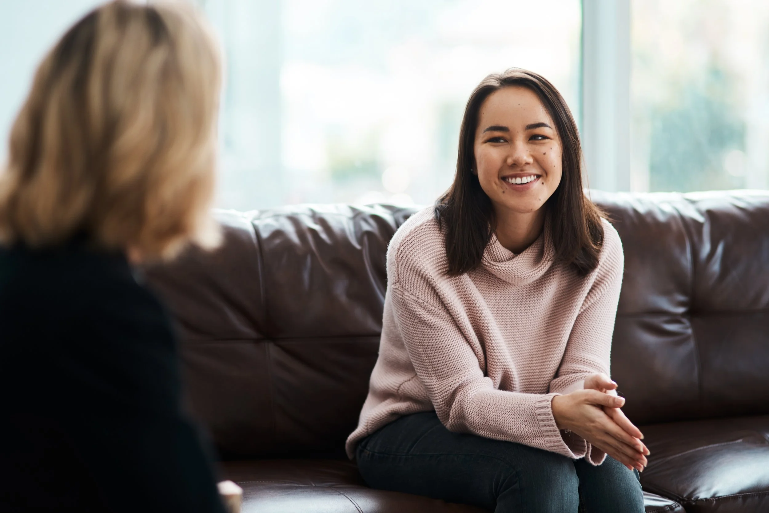 A young woman with dark hair and freckles smiling while sitting on a brown leather couch, engaged in conversation with another person whose shoulder-length blonde hair is visible in the foreground. Large windows in the background let in natural light.