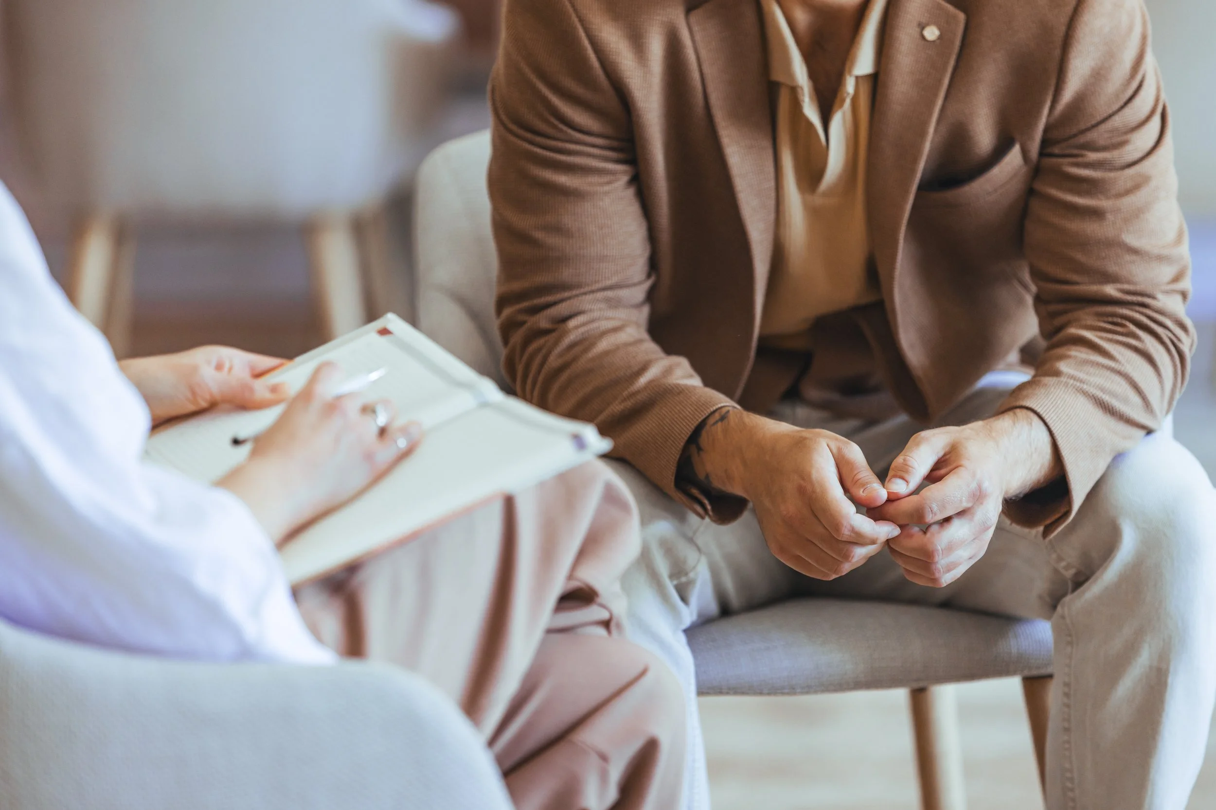 Two people having a conversation, one person holding a notepad and pen, the other sitting with hands clasped, wearing a brown blazer.