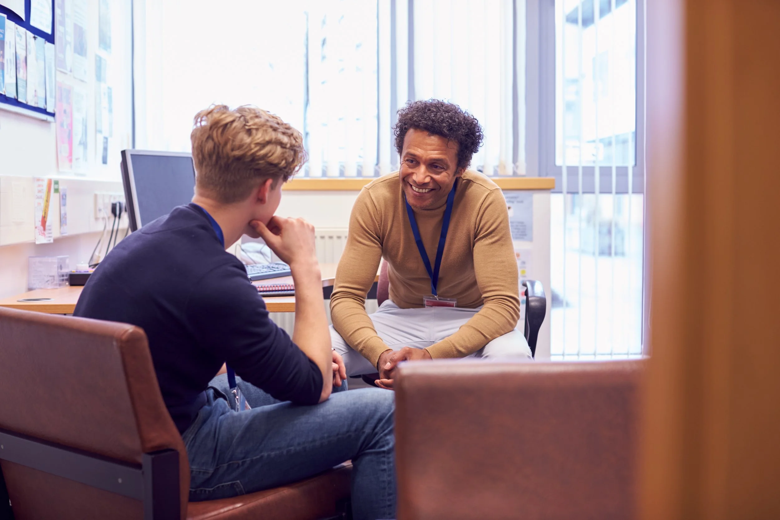 Two men having a conversation in an office, one is sitting in a brown chair with his hand on his chin, the other is sitting on a chair, smiling and leaning forward.