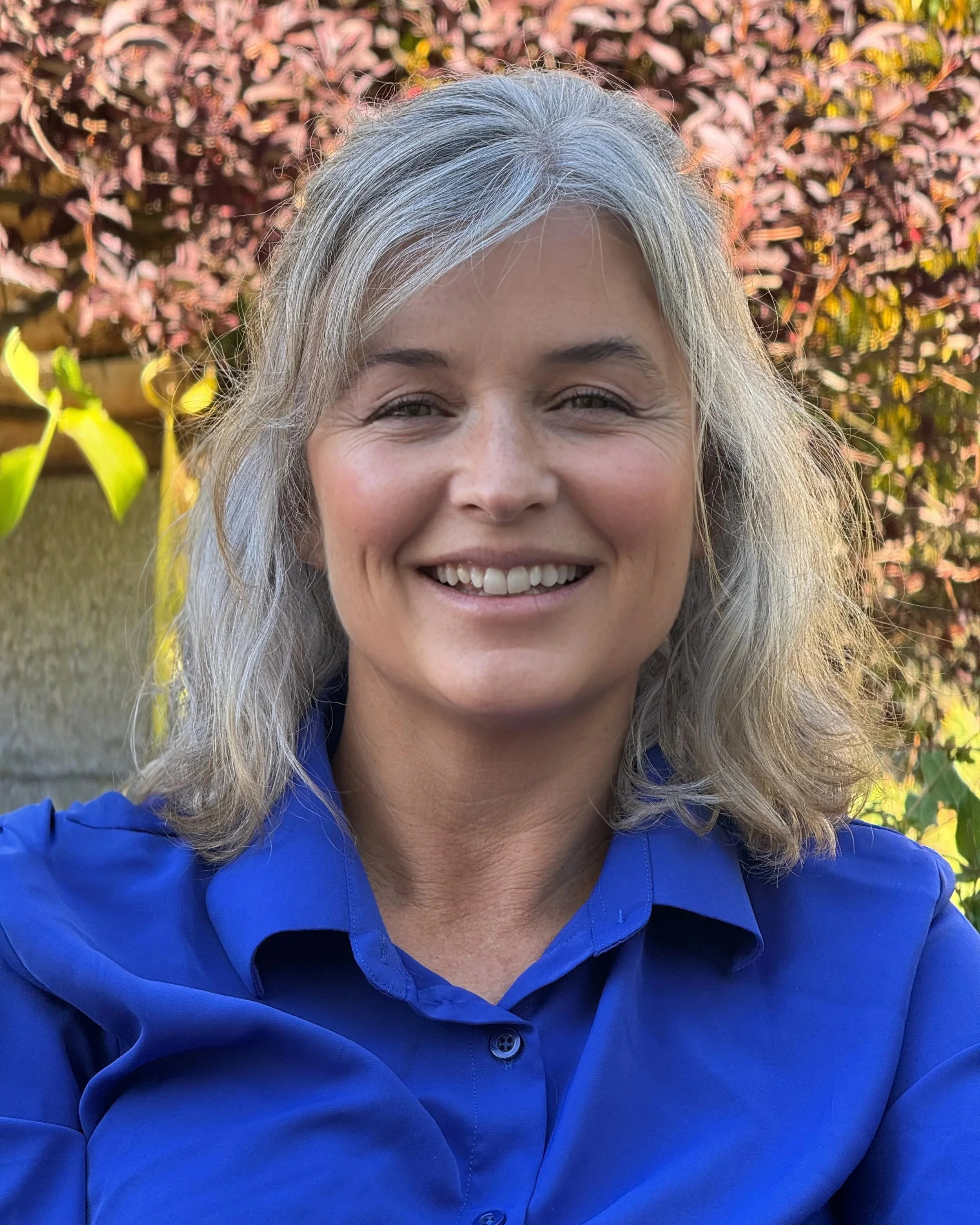 A woman with long gray hair wearing a bright blue shirt, smiling outdoors with pink and yellow foliage in the background.