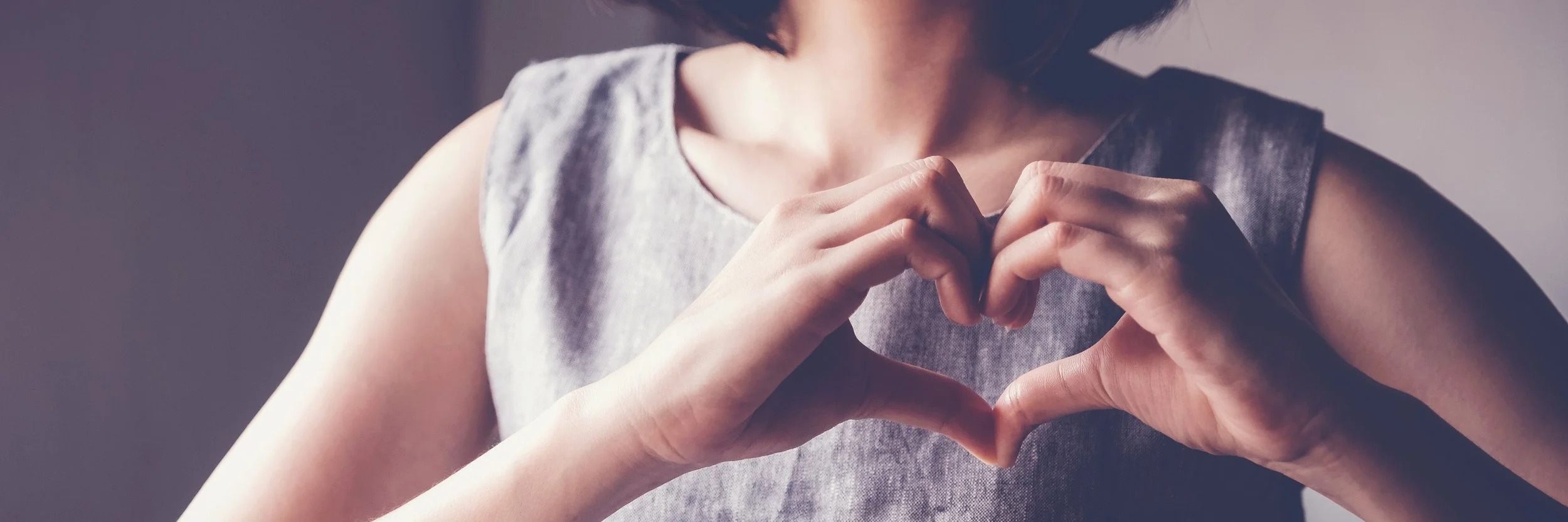 A woman wearing a sleeveless gray top making a heart shape with her hands.