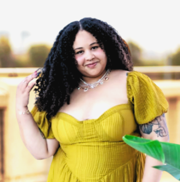 Young woman with curly hair in a yellow dress standing outdoors, smiling, with blurred background and partial view of a green object in the foreground.