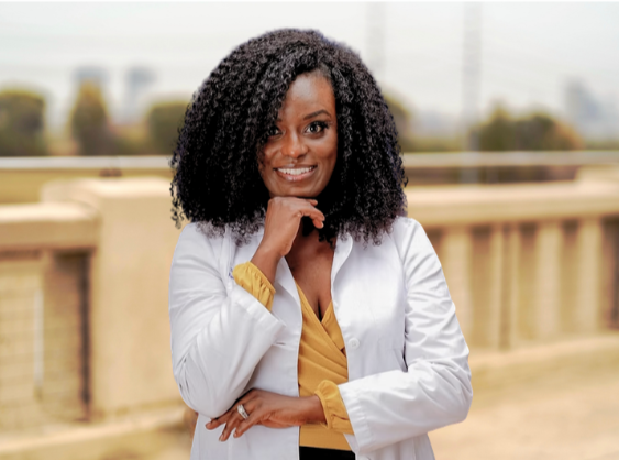 A smiling woman with curly black hair, wearing a white blazer and a yellow top, posing outdoors with a bridge in the background.