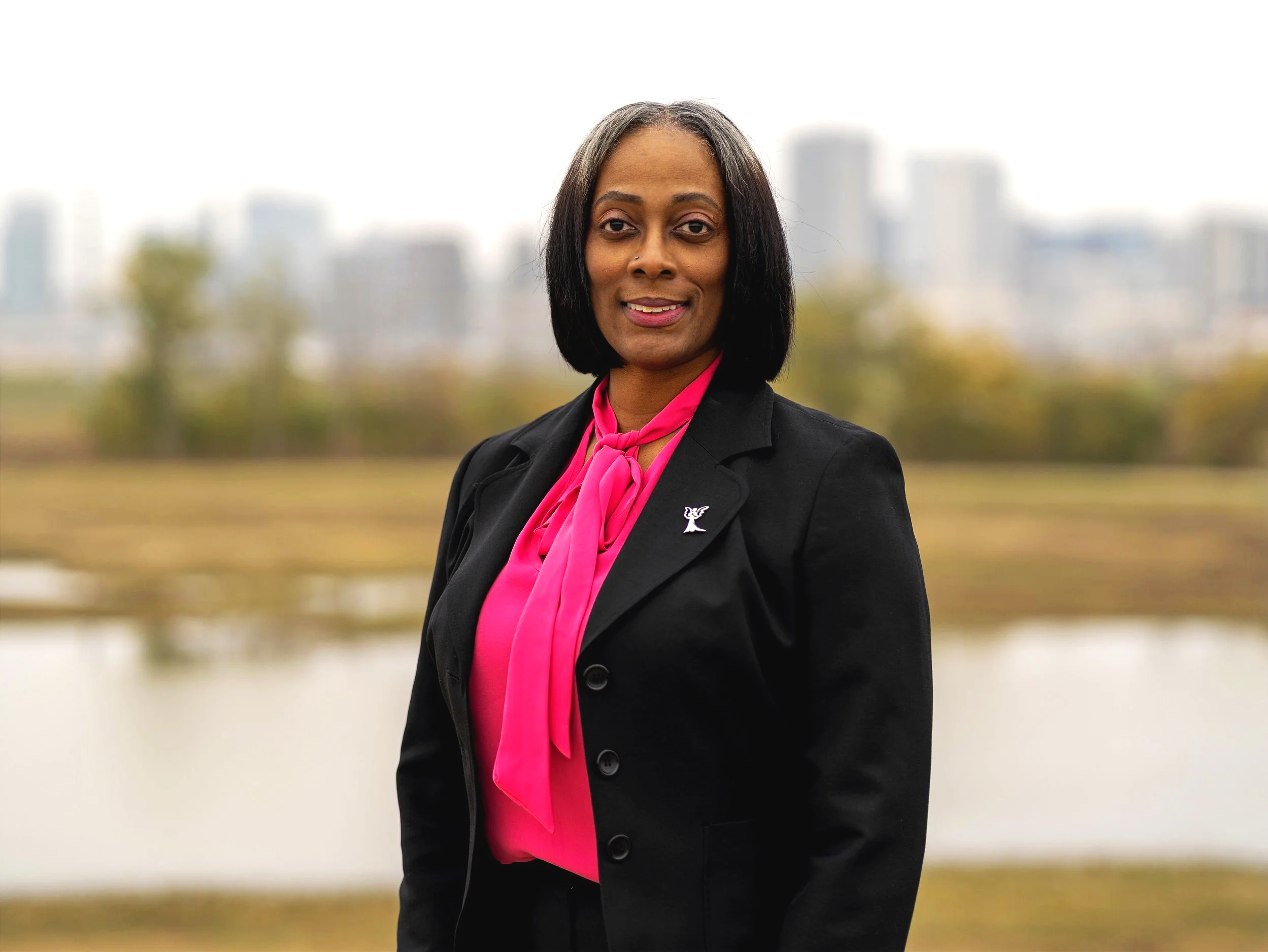 Portrait of a woman in a professional outfit standing outdoors with a blurred cityscape and water in the background.