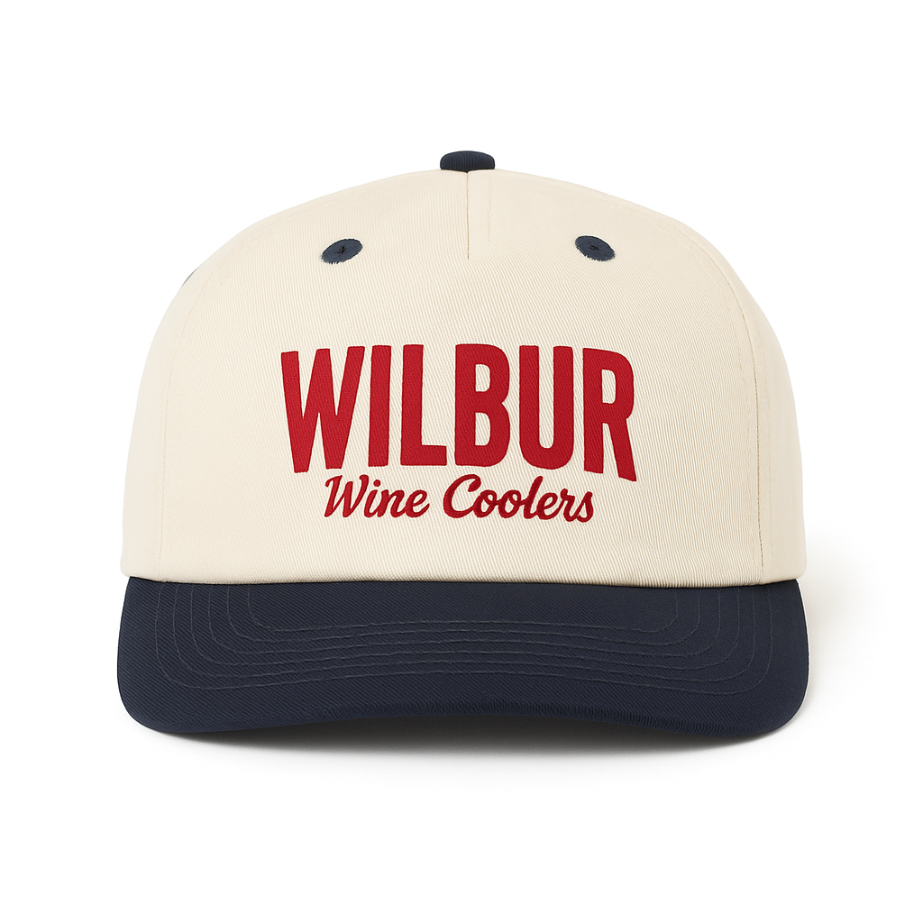 A snapback hat with an off white crown, navy brim, and navy button on top, featuring red embroidered text that says "WILBUR" and smaller red text underneath that says "Wine Coolers".