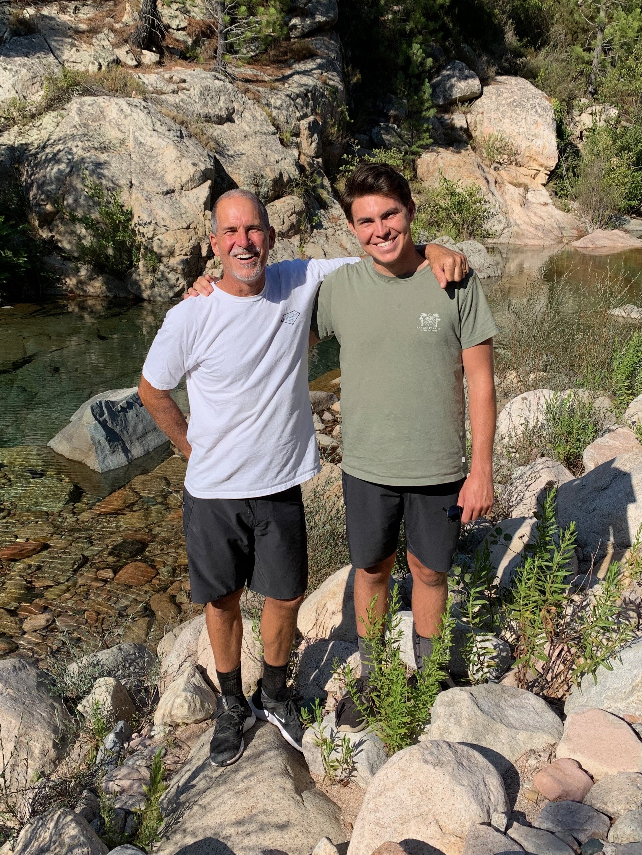 Two men, one older and one younger, standing on rocks by a rocky creek, smiling, with arms around each other's shoulders on a sunny day in a natural outdoor setting.