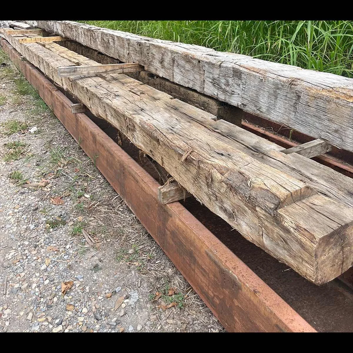 Stacked antique reclaimed hand-hewn timbers with visible tool marks, aged patina, and weathered surfaces stored outdoors.