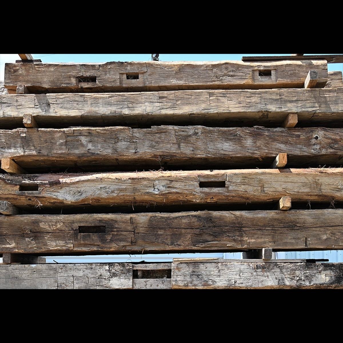 Stacked antique reclaimed hand-hewn timbers with visible tool marks, mortise pockets, and aged weathered surfaces.