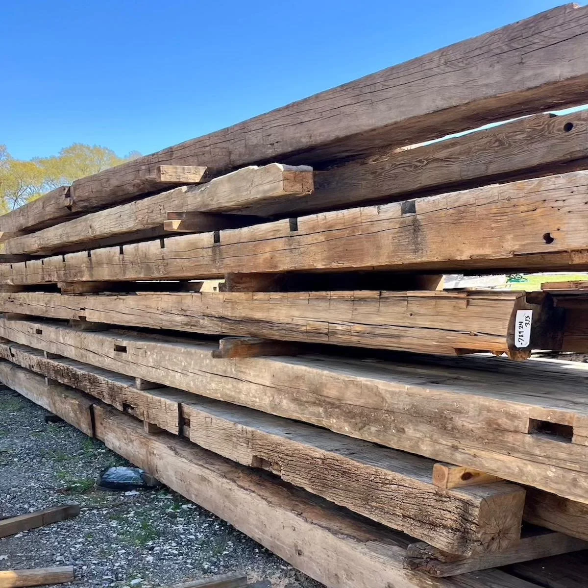 Stacked antique reclaimed hand-hewn timbers, showing tool marks, weathered patina, and different lengths.