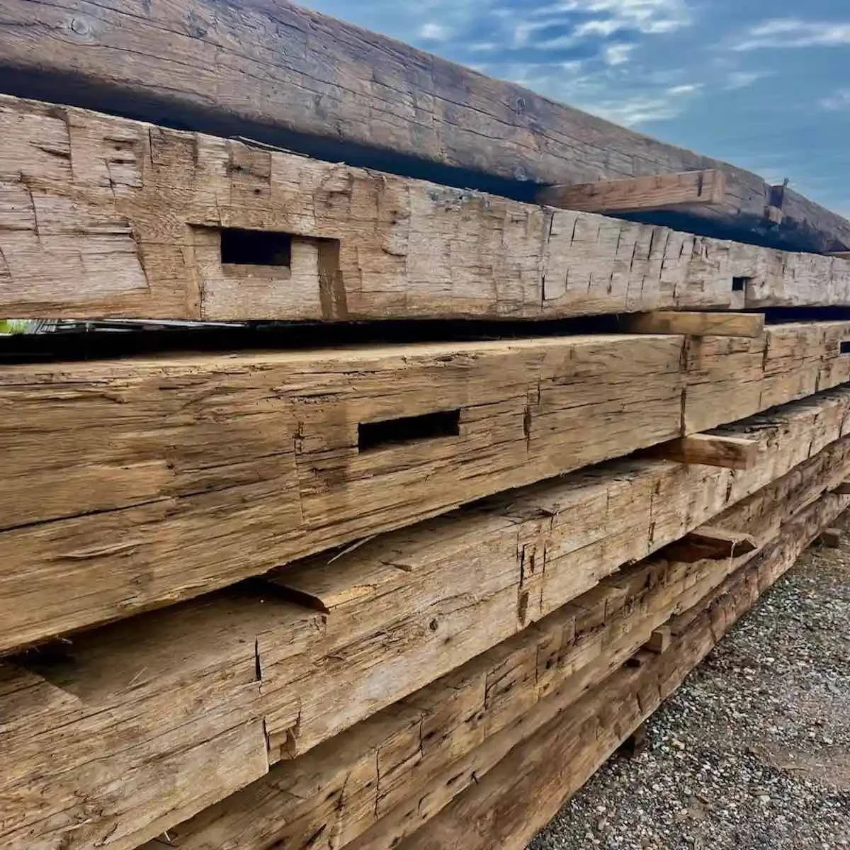 Antique reclaimed hand-hewn timbers with visible mortise pockets, stacked outdoors with aged patina.