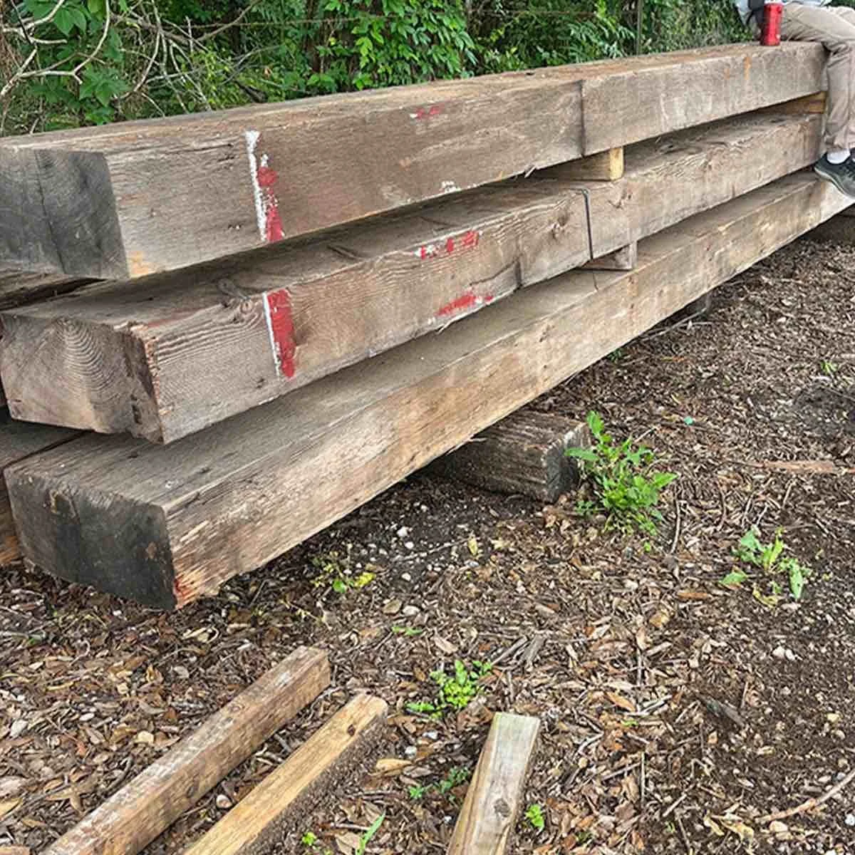 Antique reclaimed industrial timbers with weathered surfaces and remnants of red paint, stacked outdoors showing aged patina and rustic character.