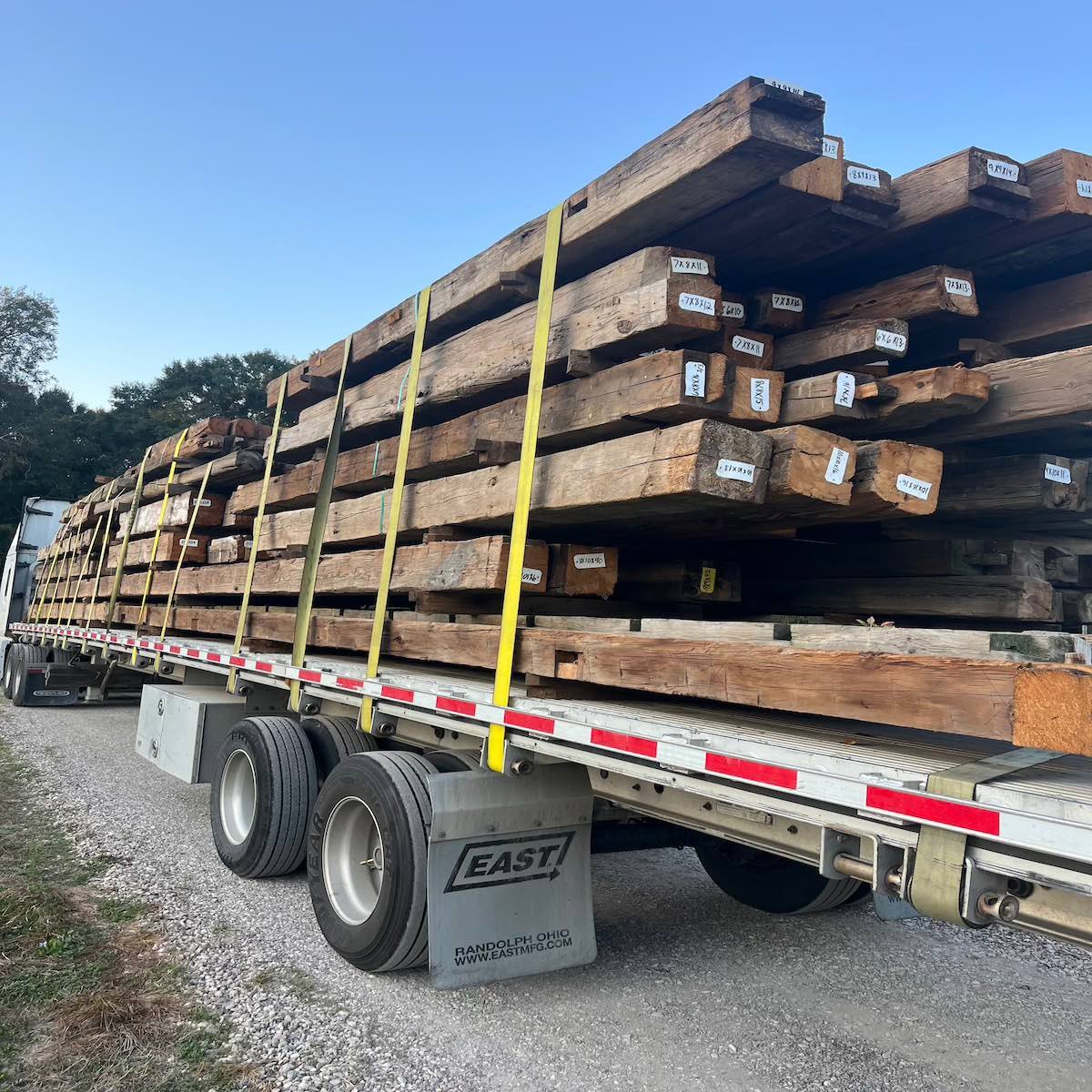 Antique reclaimed hand-hewn timbers loaded and strapped on a flatbed trailer for transport, showing timbers with dimension tags.