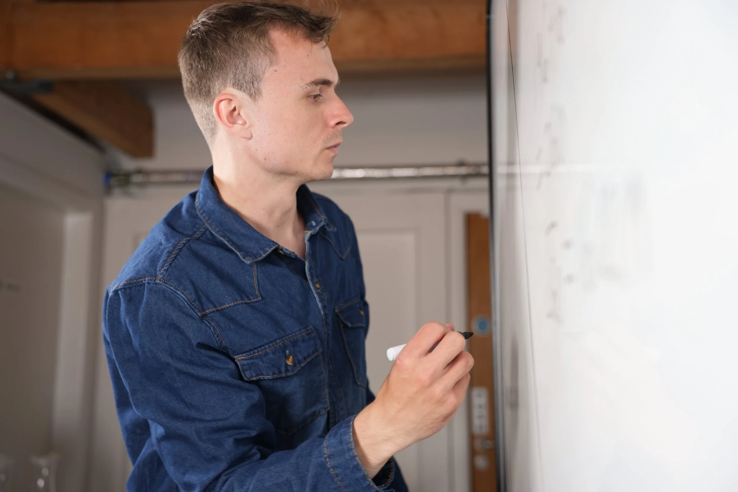 A young man in a denim jacket writing on a whiteboard in a room with a wooden ceiling.