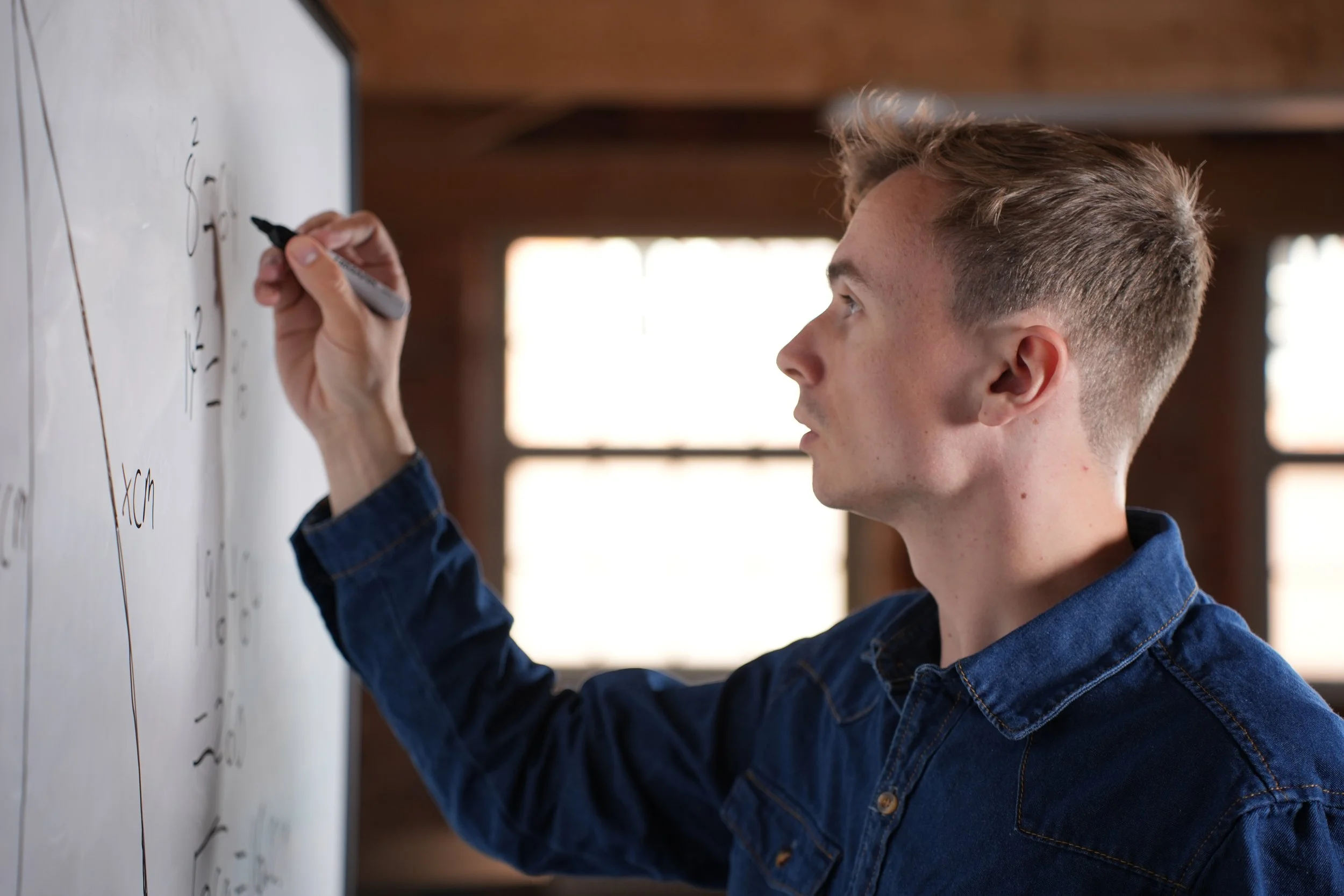 A young man writing on a whiteboard with a black marker, with his profile facing the right side of the image, in a room with large windows.