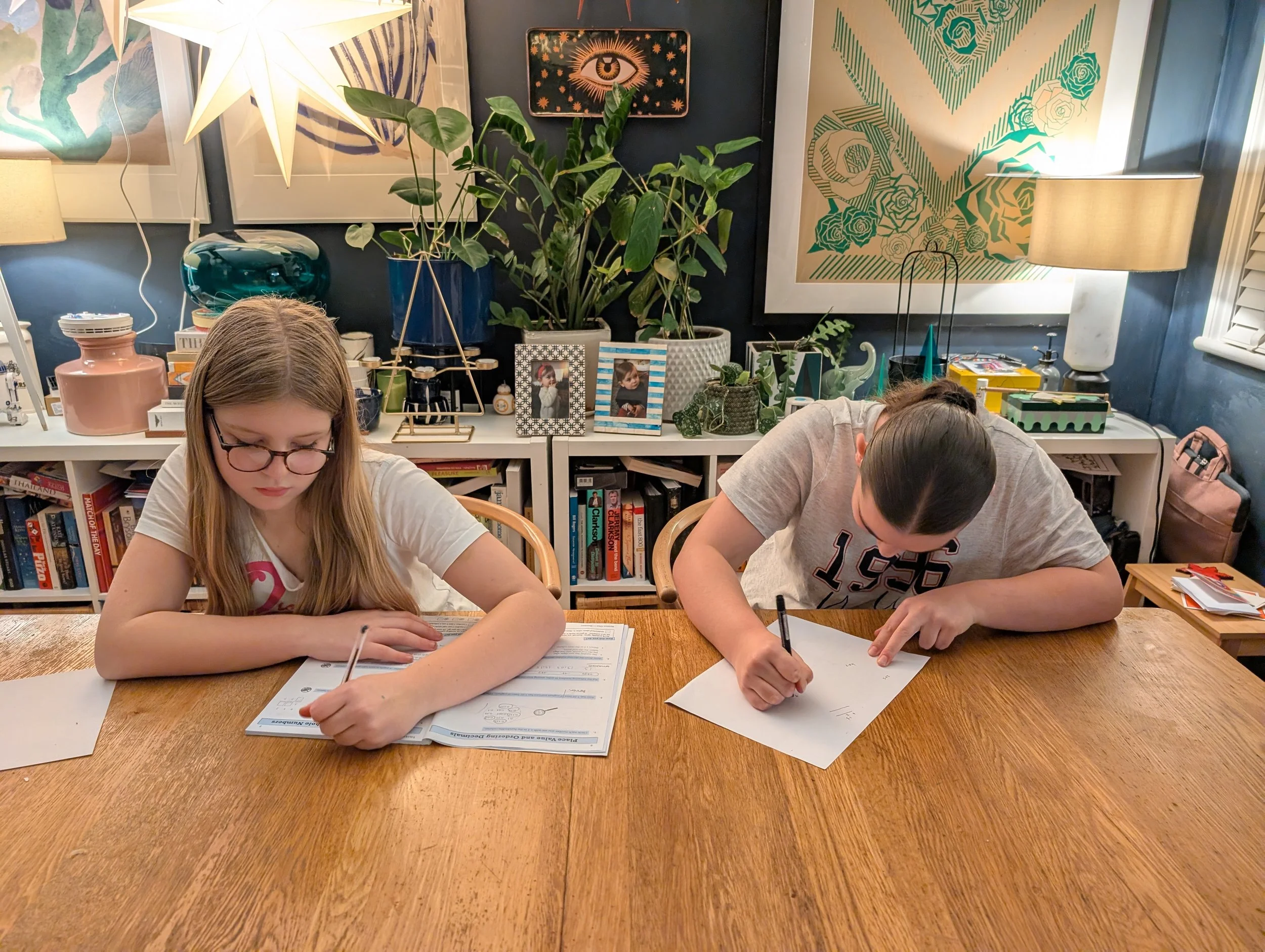 Two girls sitting at a wooden table, doing homework or drawing. The girl on the left has long blonde hair and glasses, and the girl on the right has dark hair pulled back. The background features bookshelves, colorful artwork, plants, and decorative lamps.