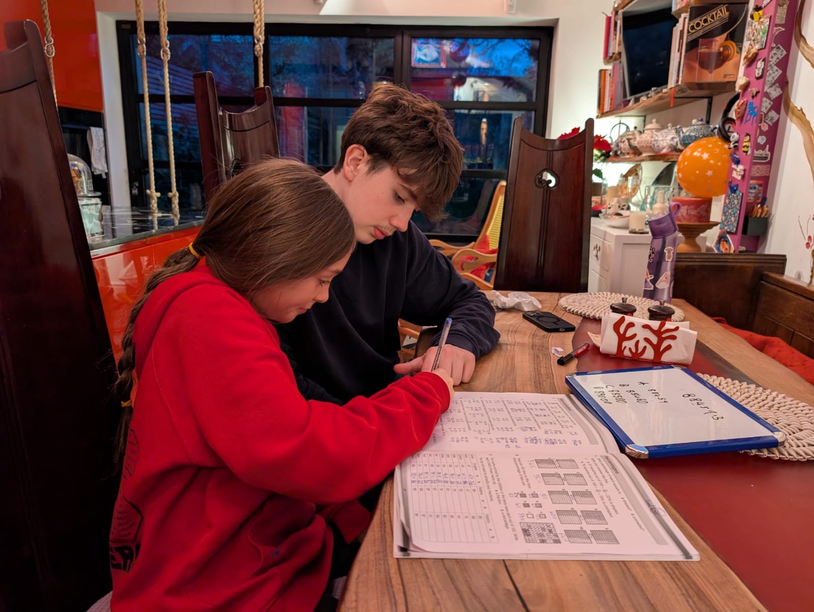 A girl and a boy working on homework at a wooden dining table, surrounded by holiday decorations, including a gift box with red reindeer antlers and a whiteboard with math problems.