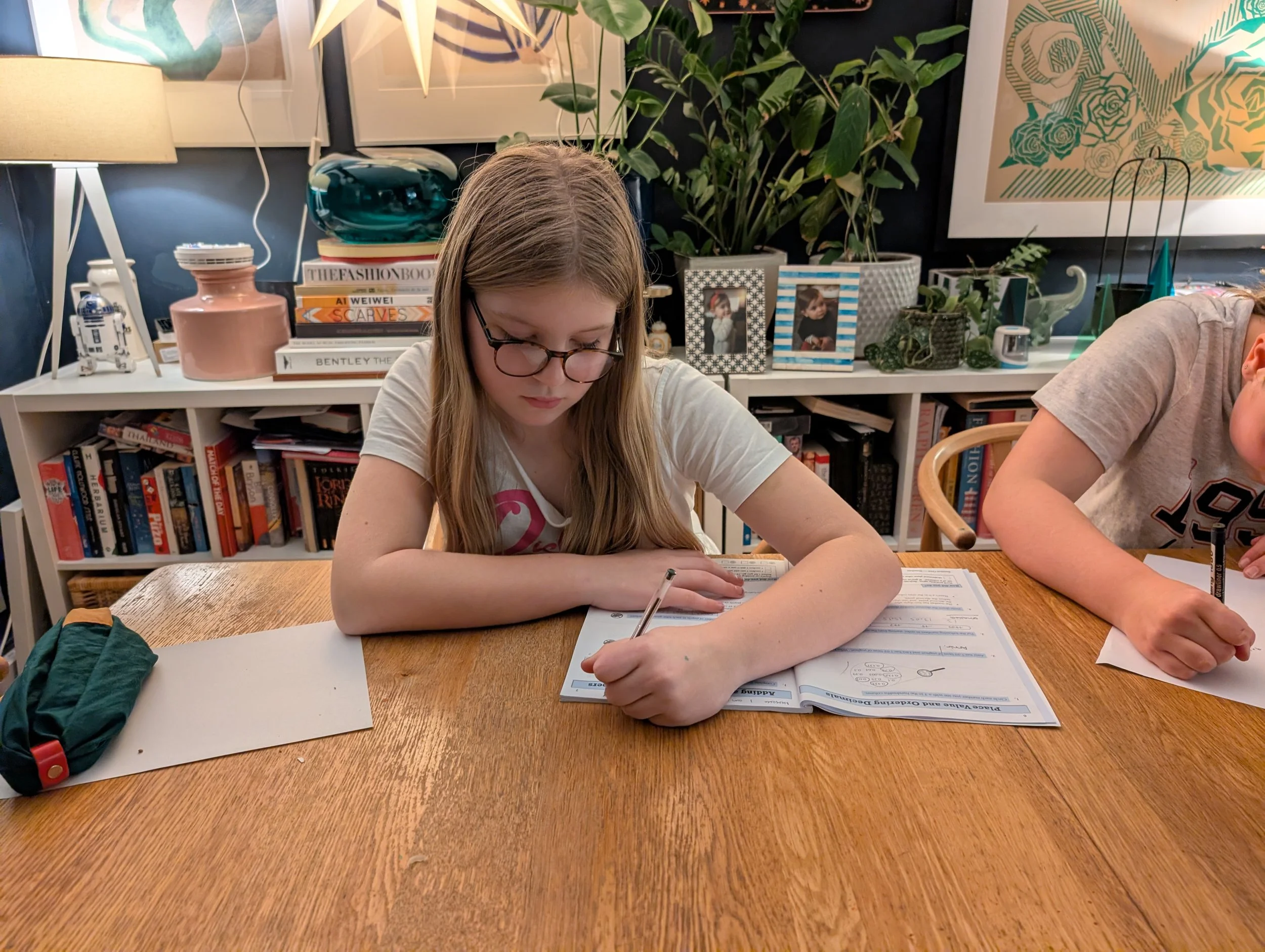 A young girl with glasses sitting at a wooden table, working on a worksheet with a pencil. There is a school backpack on the left side of the table and another student partially visible on the right. The background shows a bookshelf, framed pictures, decorative objects, and plants.