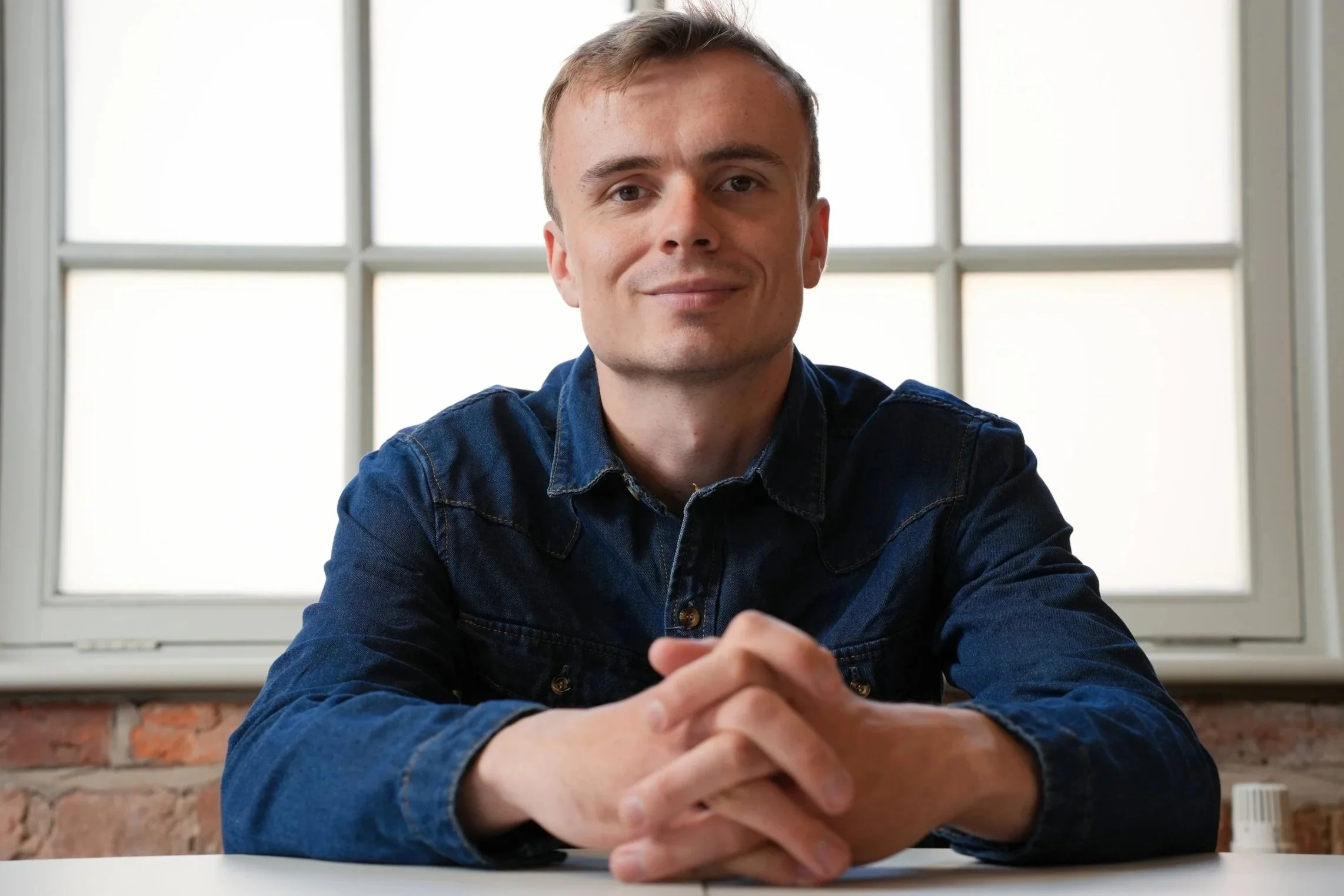 A young man with short light brown hair, wearing a denim shirt, sitting at a table with hands clasped, smiling, in front of a window with multiple panes in a bright room.