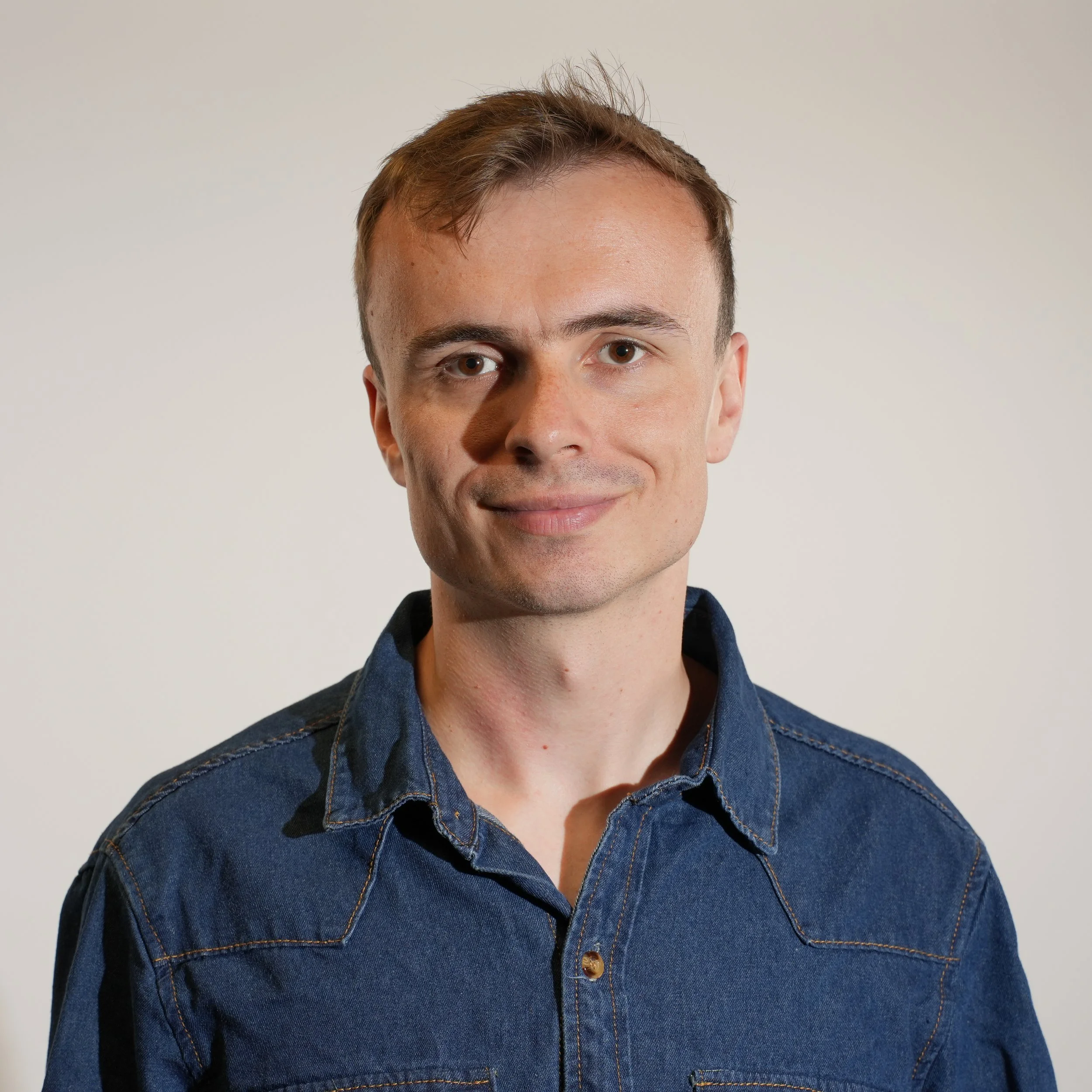Portrait of a young man with short brown hair, wearing a denim shirt, smiling softly, against a plain light background.