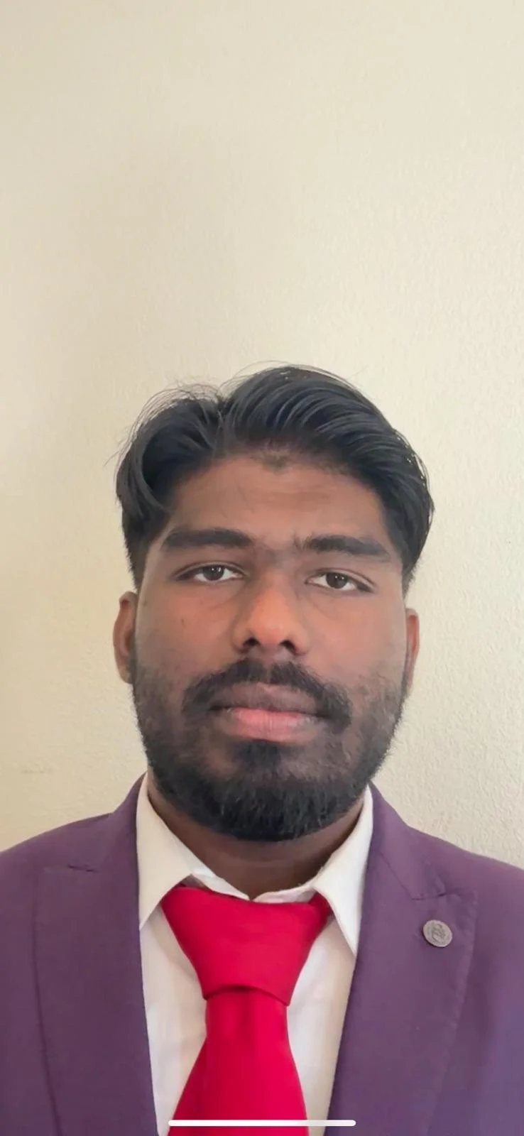 Portrait of a man with dark hair and beard, wearing a purple suit, white shirt, and red tie, standing against a plain beige wall.