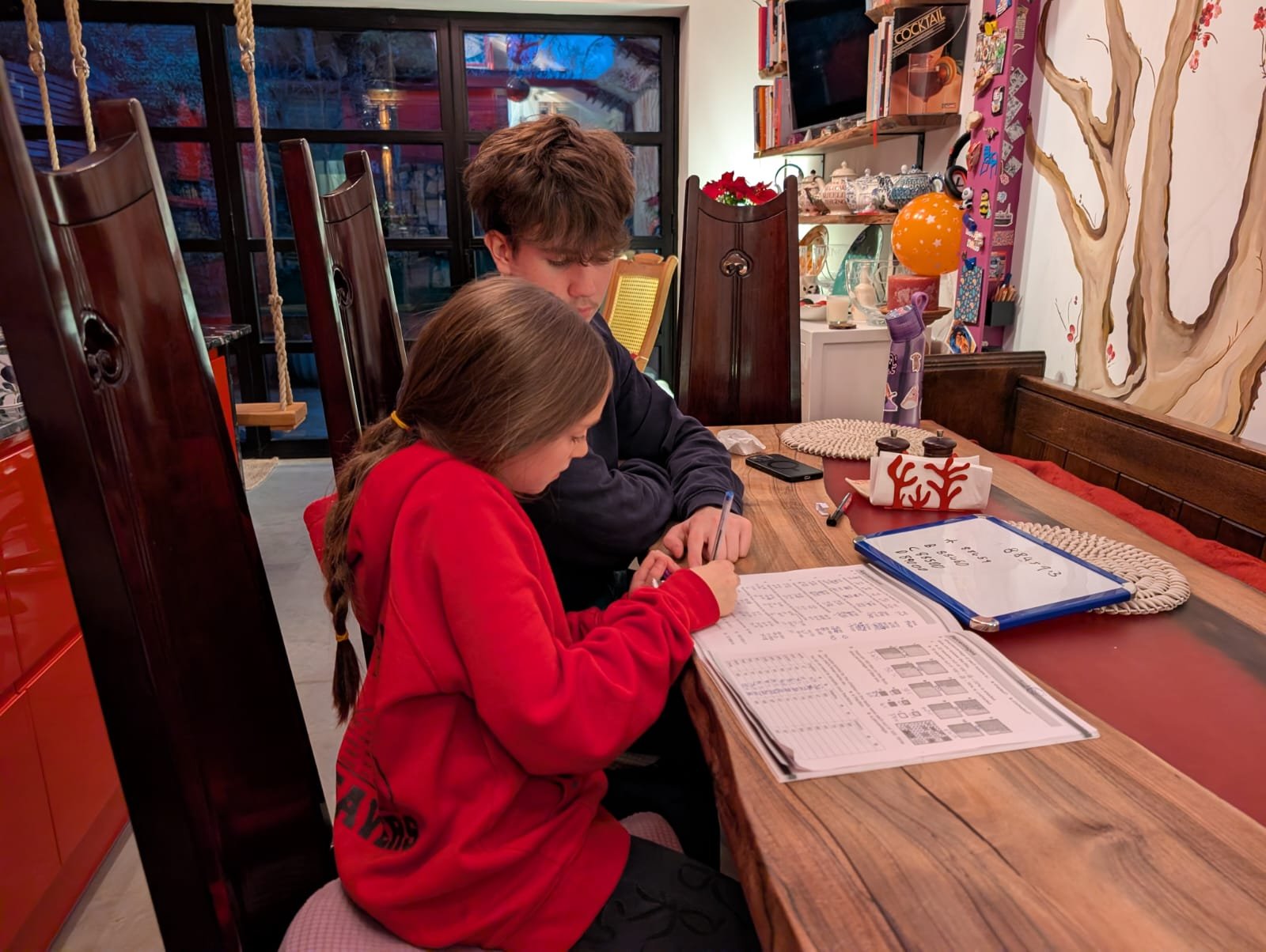 Two children, a girl in a red sweatshirt and a boy in a black hoodie, sitting at a wooden table working on homework with papers and pens, in a decorated room with books, toys, and a mural of a tree on the wall.