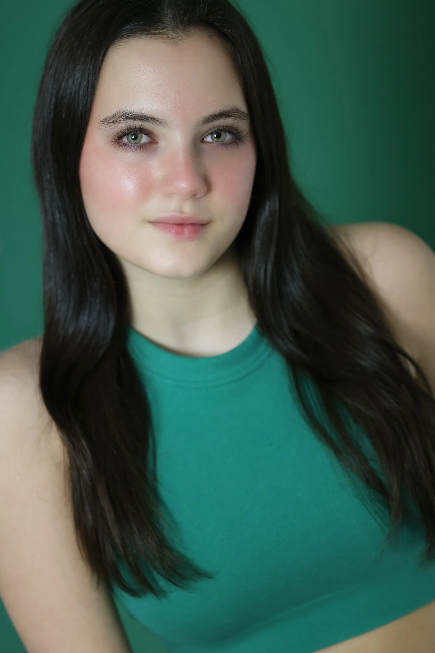 Close-up of a young woman with long dark hair, green eyes, and light skin, wearing a green sleeveless top against a green background.