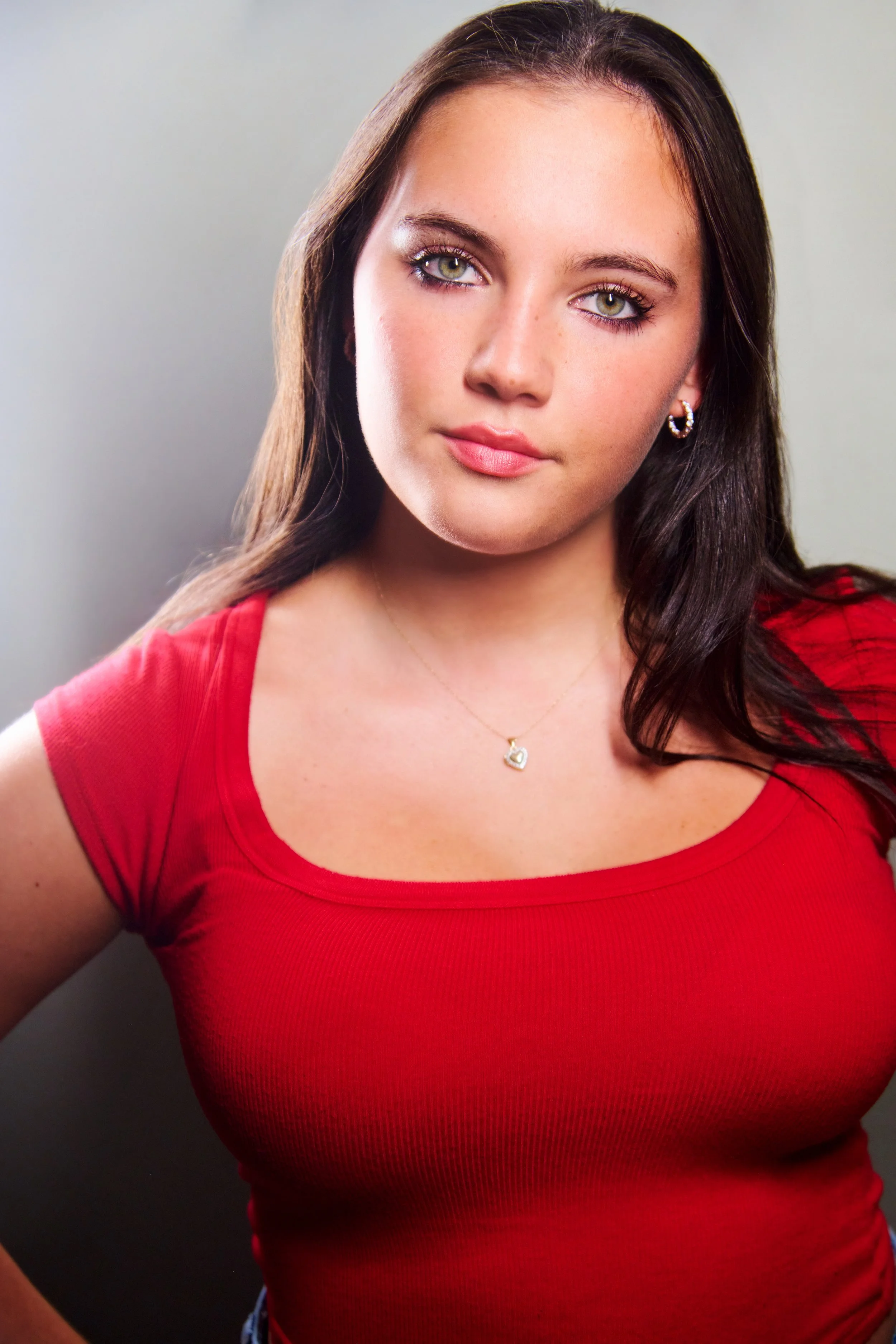 A young woman with long brown hair, wearing a red top, silver earrings, and a necklace with a heart-shaped pendant.
