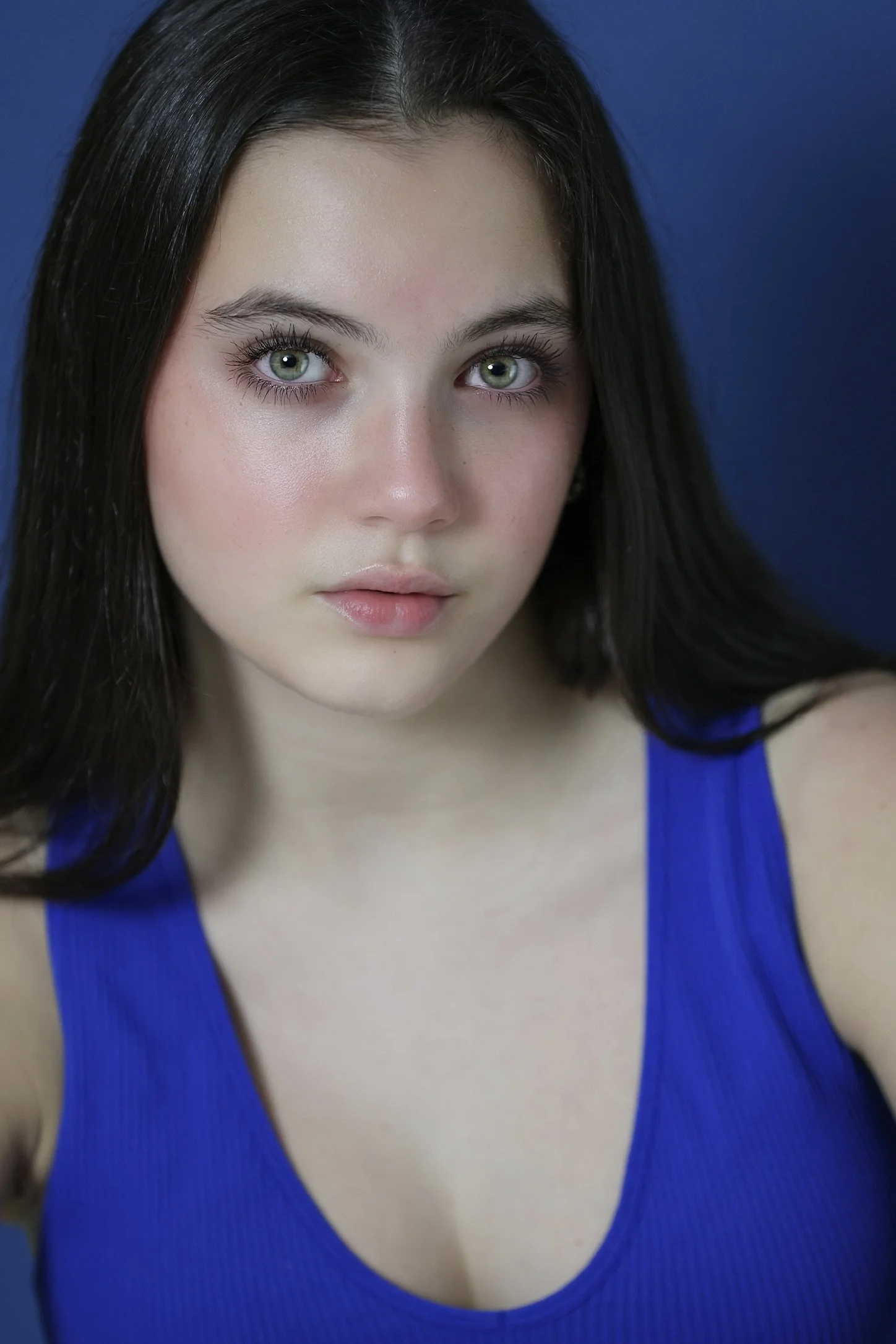 Close-up portrait of a young woman with long dark hair, green eyes, and fair skin, wearing a blue top, against a dark blue background.