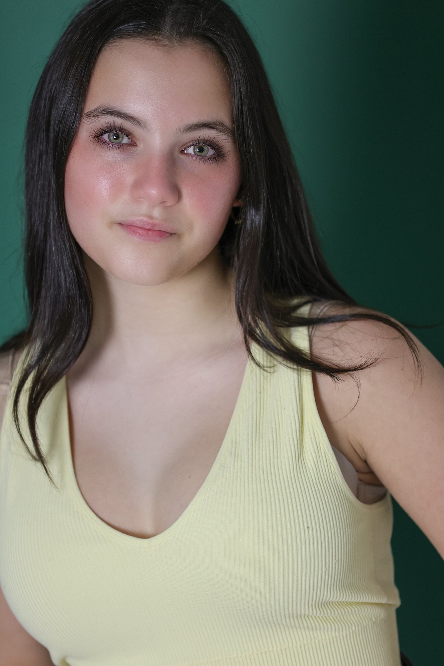 Close-up of a young woman with long dark hair and green eyes wearing a yellow sleeveless top against a green background.
