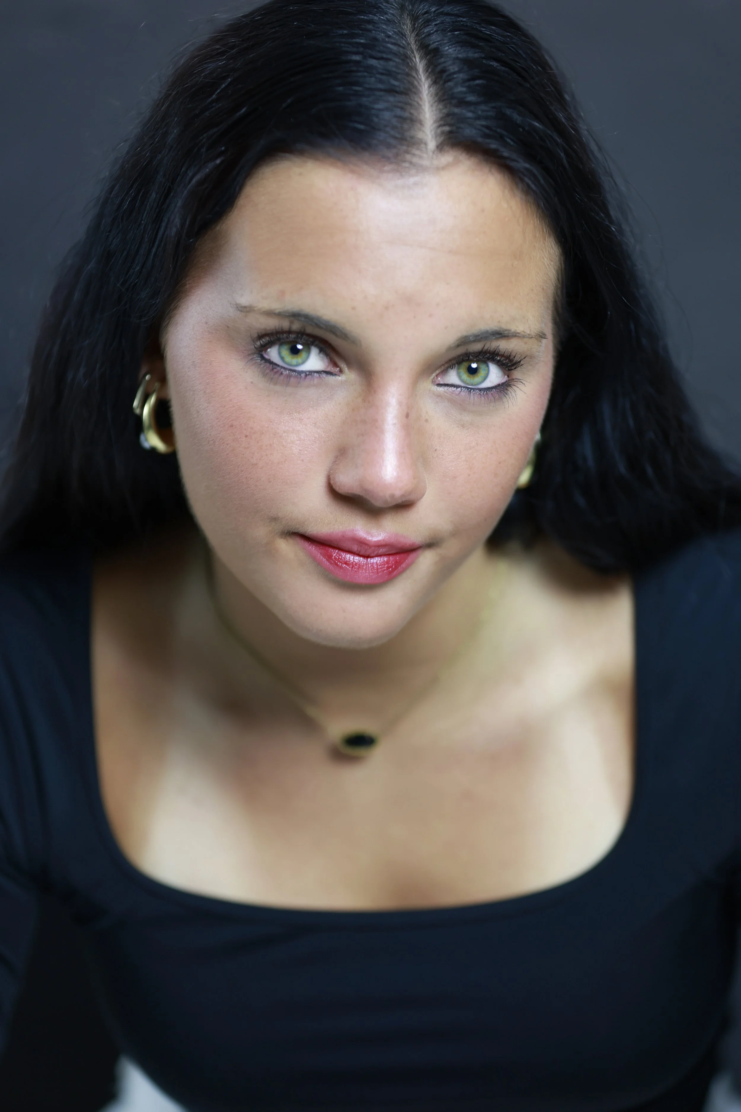 Close-up of a young woman with long dark hair, fair skin, and striking green eyes, wearing a red top, looking directly at the camera against a neutral background.