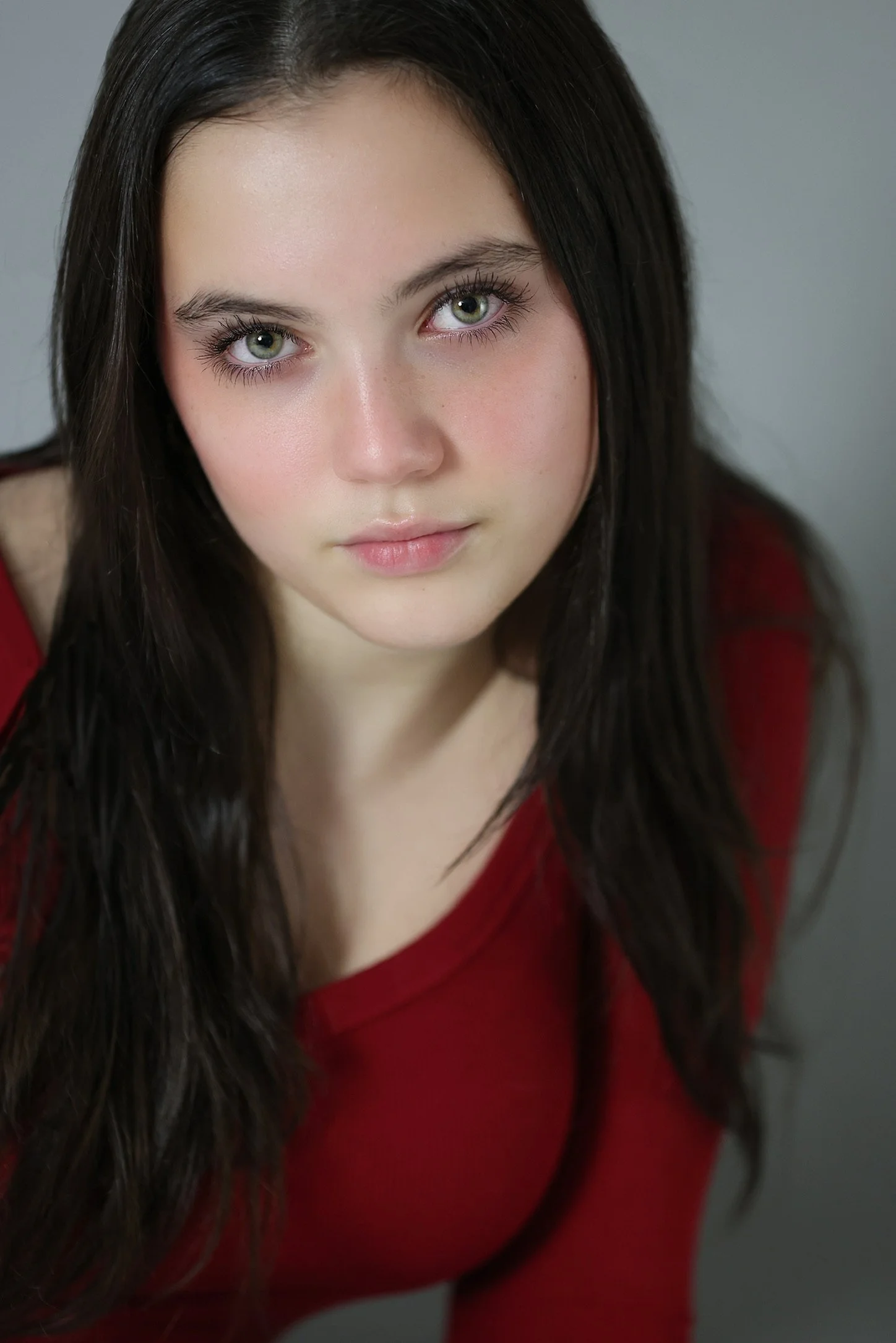 Close-up of a young woman with long dark hair, fair skin, and striking green eyes, wearing a red top, looking directly at the camera against a neutral background.