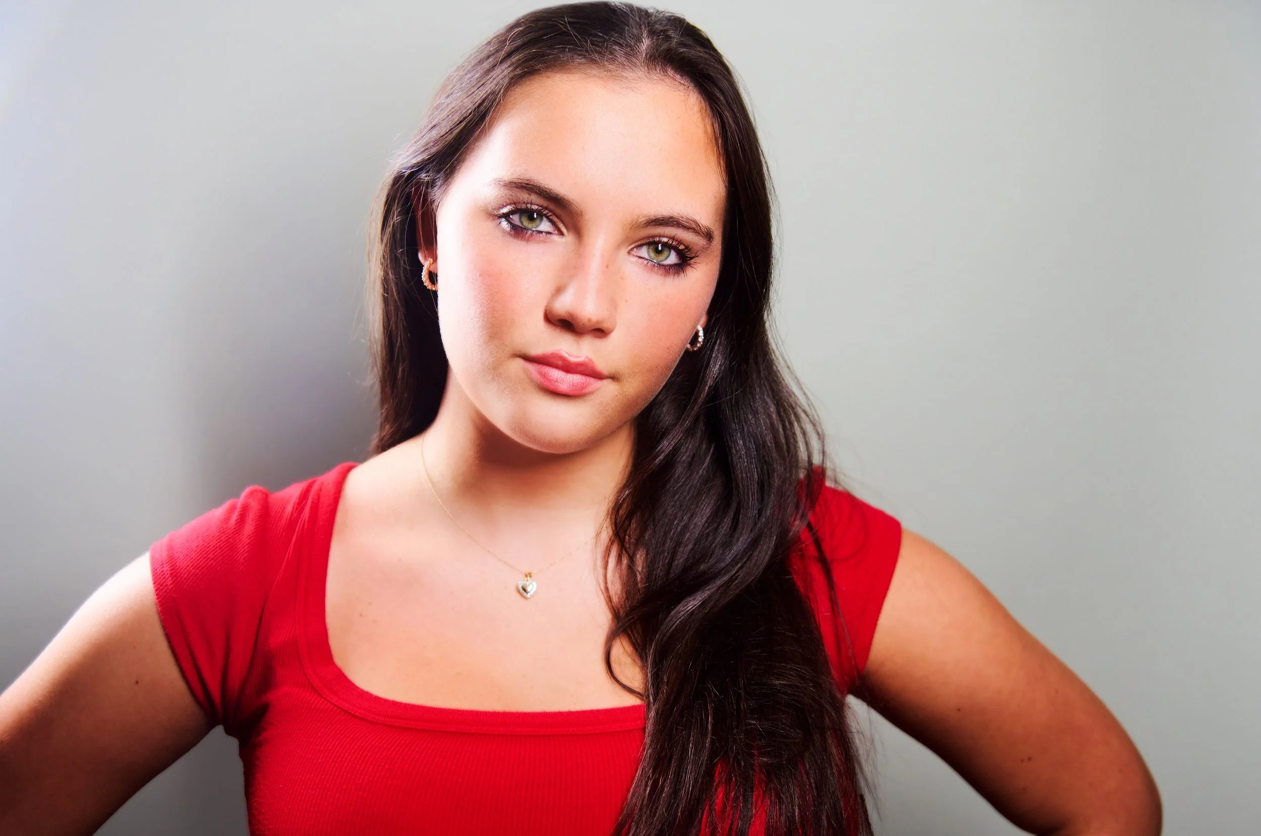 A young woman with long dark hair, wearing a red top and heart-shaped necklace, standing against a neutral background.