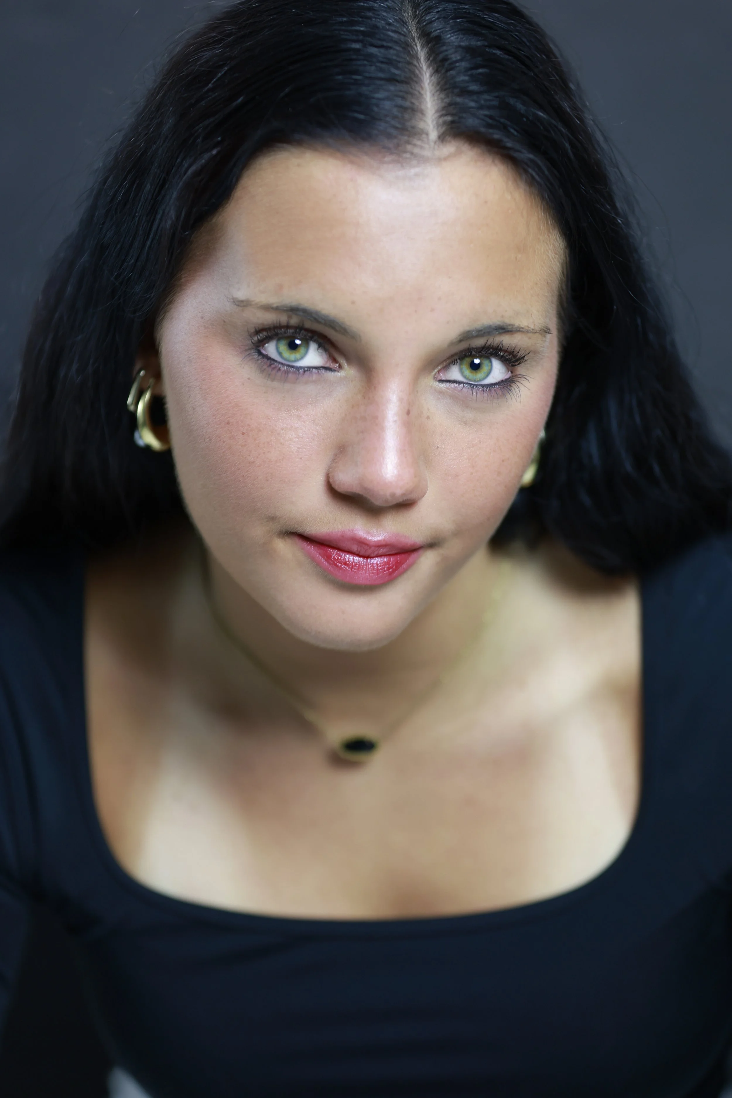 Close-up of a young woman with long dark hair, green eyes, and light skin, wearing a green sleeveless top against a green background.