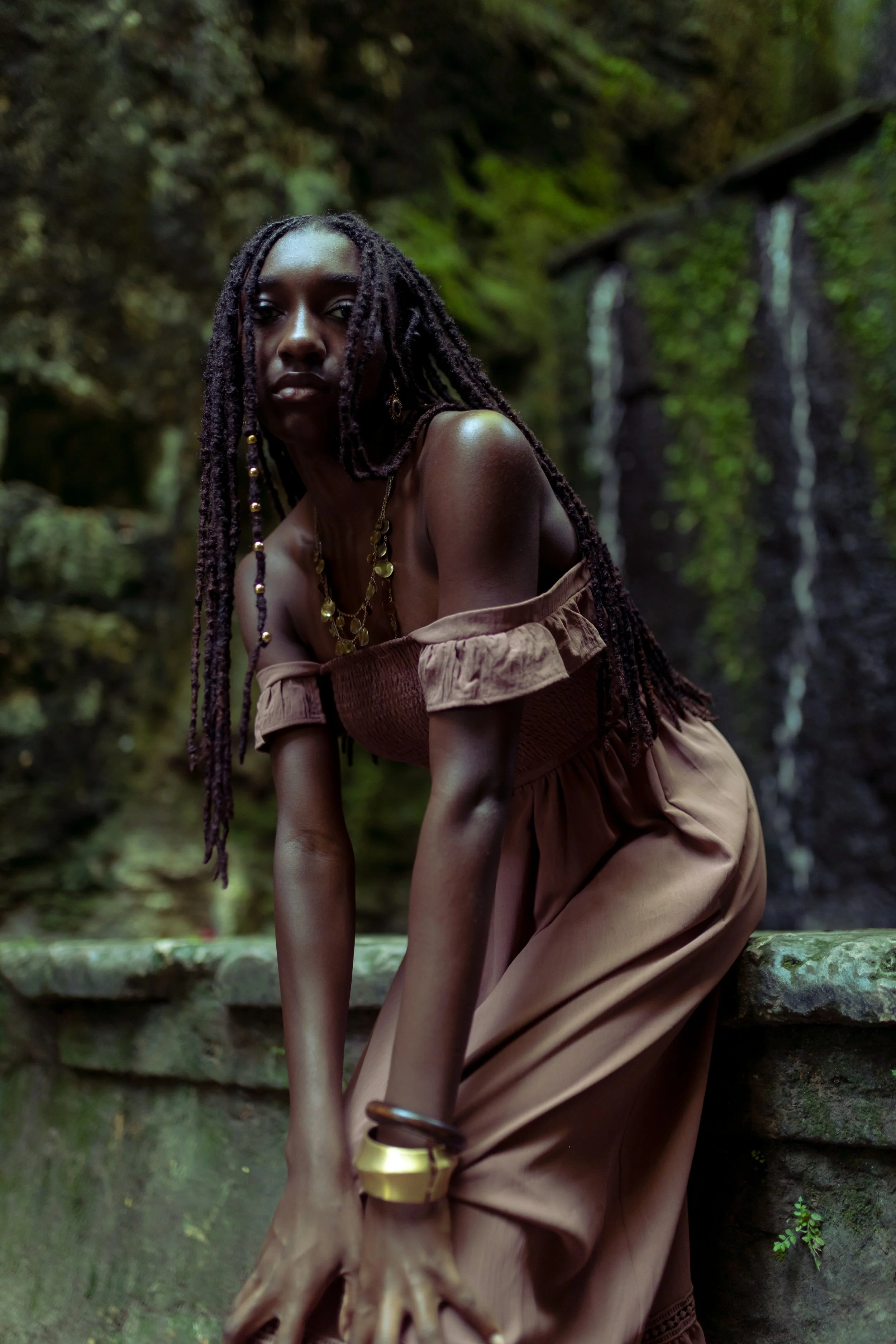 A woman with long, twisted hair wearing a brown off-shoulder dress and gold jewelry, standing outdoors near a mossy stone wall and a waterfall.