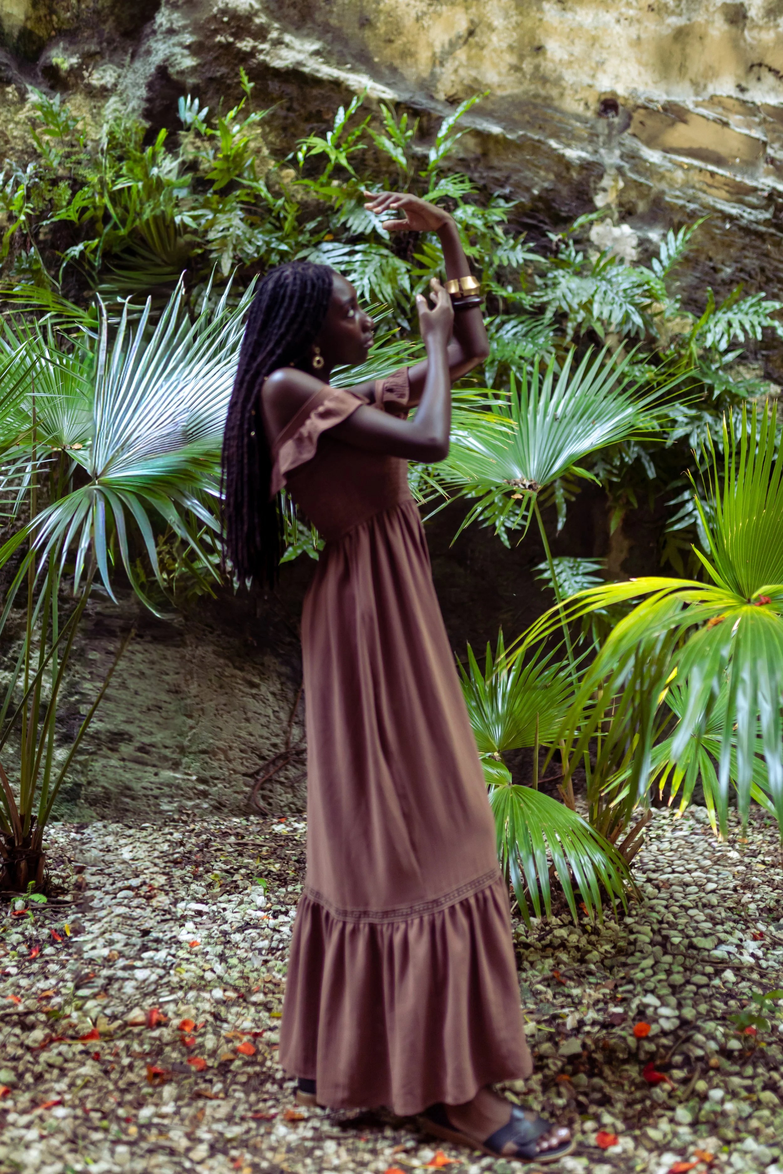 A woman with long dreadlocks wearing a brown dress standing among tropical plants, striking a graceful pose in a lush, green environment.