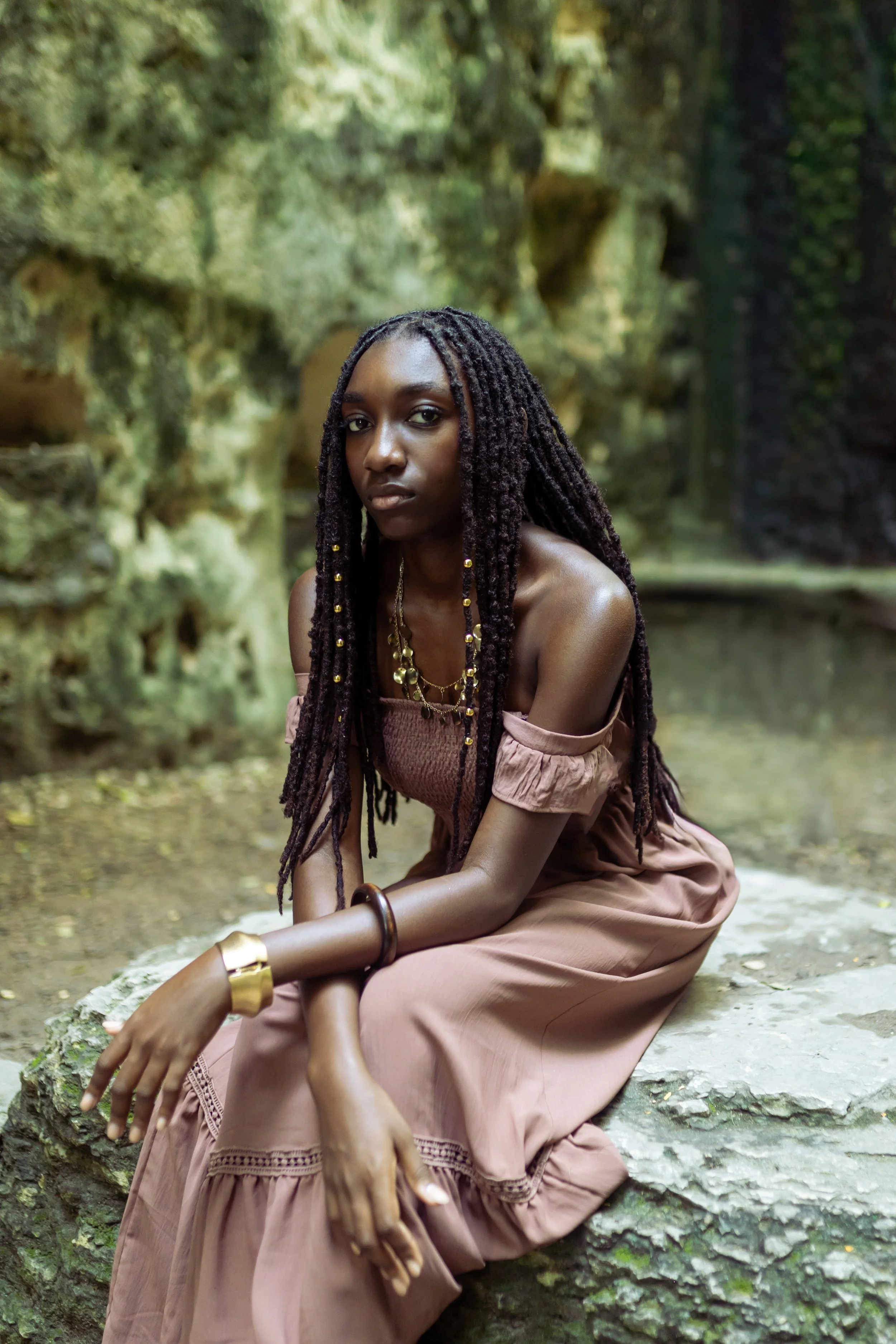 A woman with long dreadlocks and dark skin, dressed in a pink off-shoulder dress, sits on a large rock in a lush green forest, looking at the camera with a serious expression.
