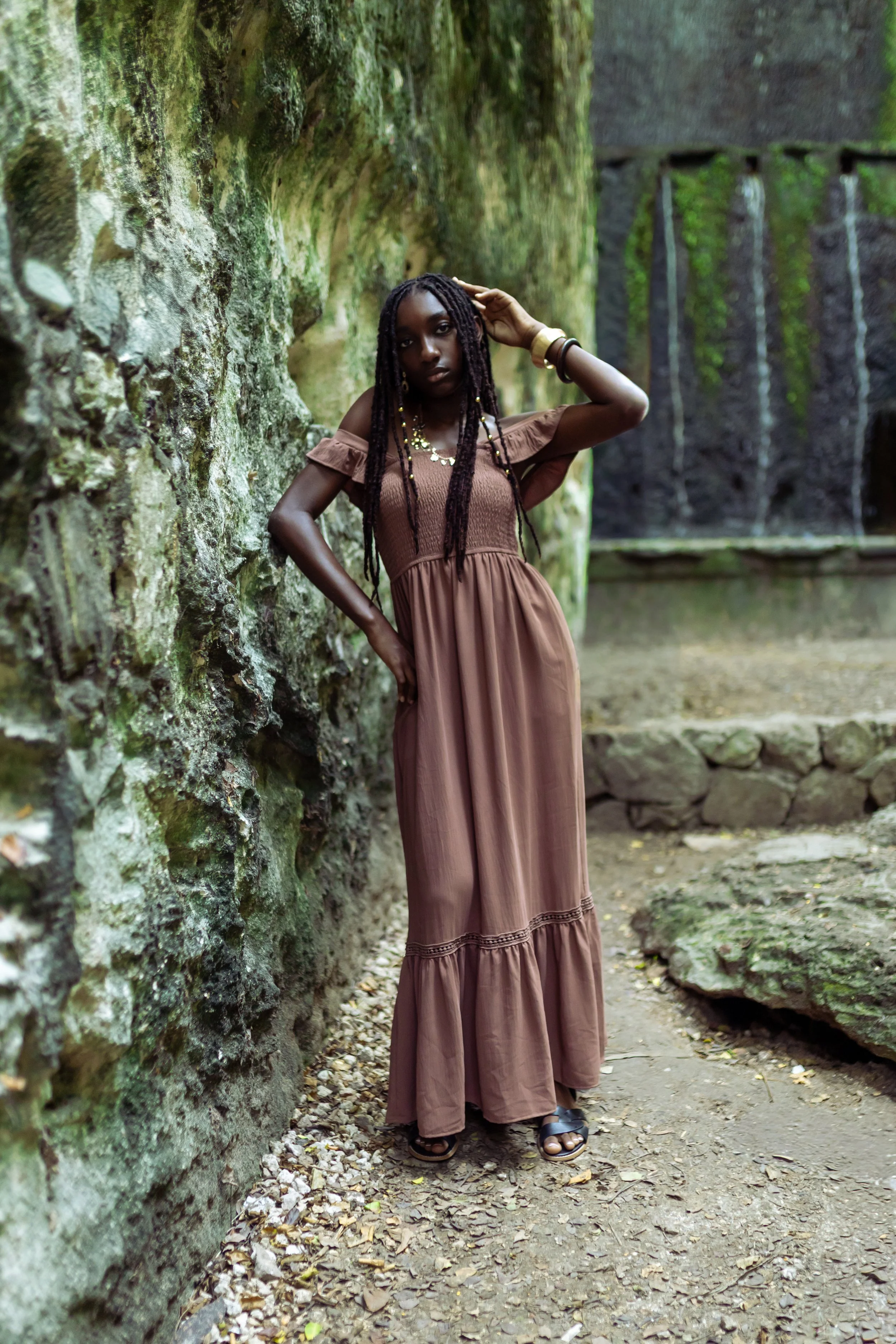 A woman with long dark braids wearing a brown off-shoulder maxi dress and sandals standing against a moss-covered stone wall outdoors, with a waterfall in the background.