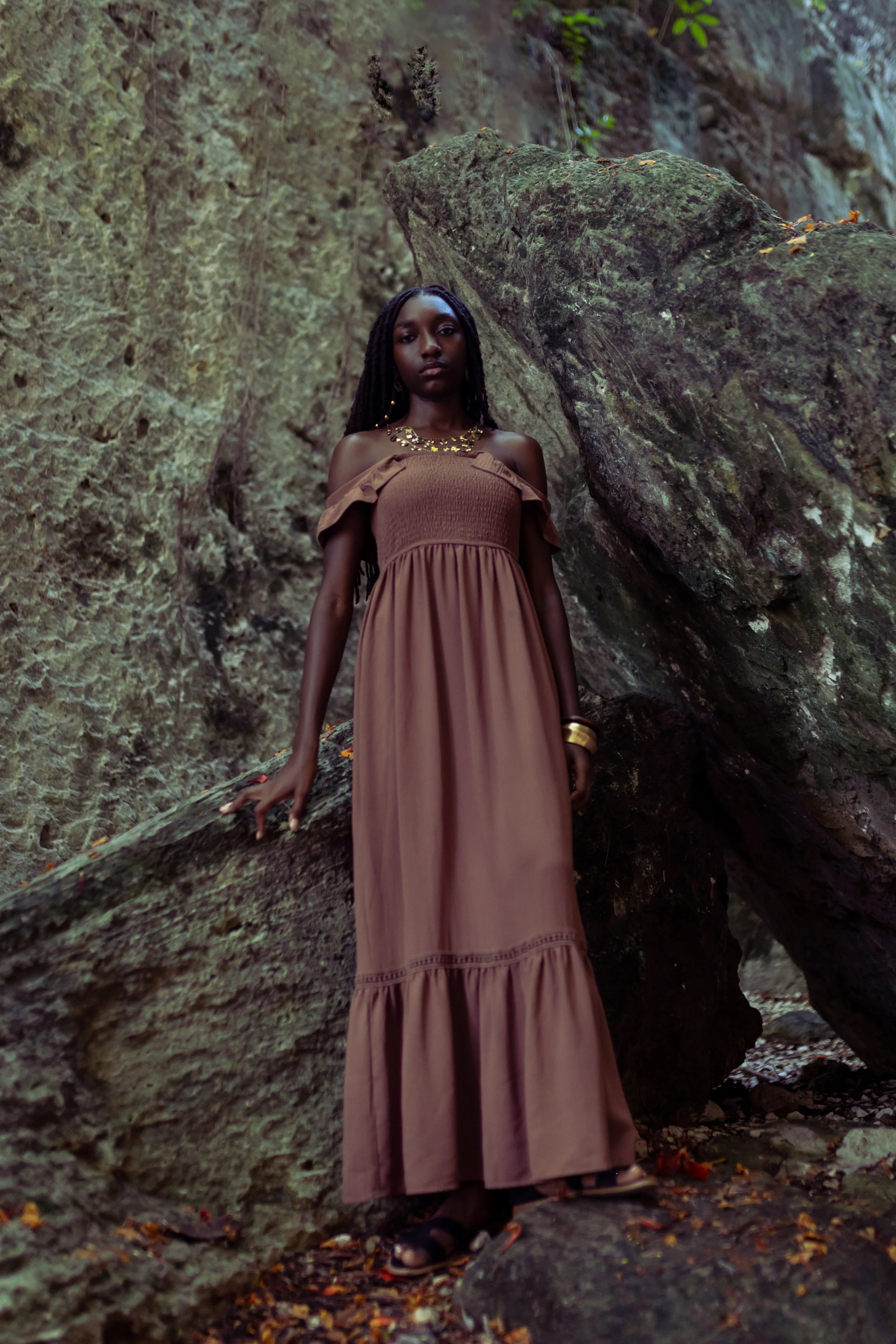 A woman in a long brown dress standing among large rocks, with trees in the background.