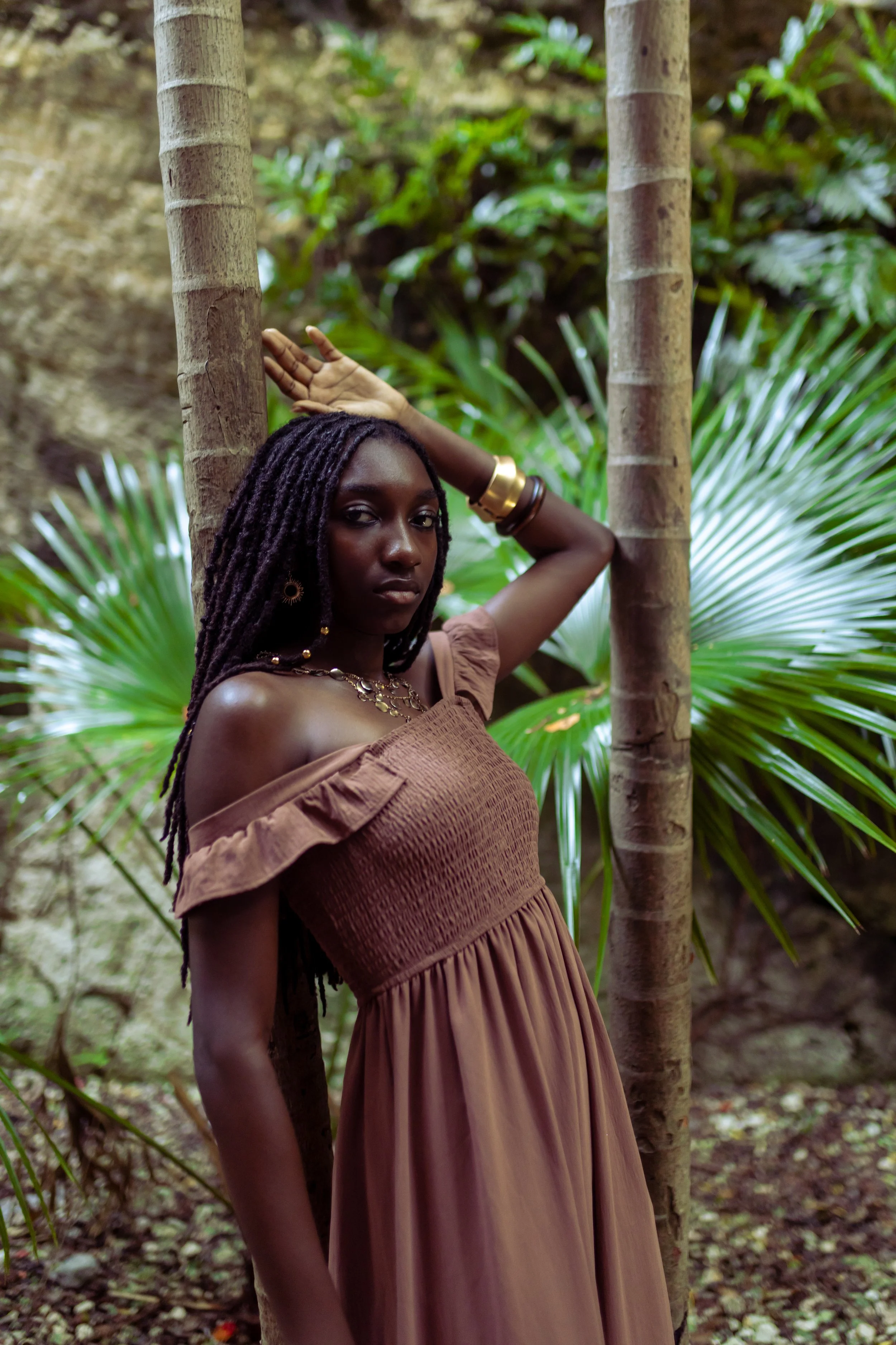 A woman with long dreadlocks and dark skin posing outdoors among tropical plants, wearing a brown off-the-shoulder dress and gold jewelry.