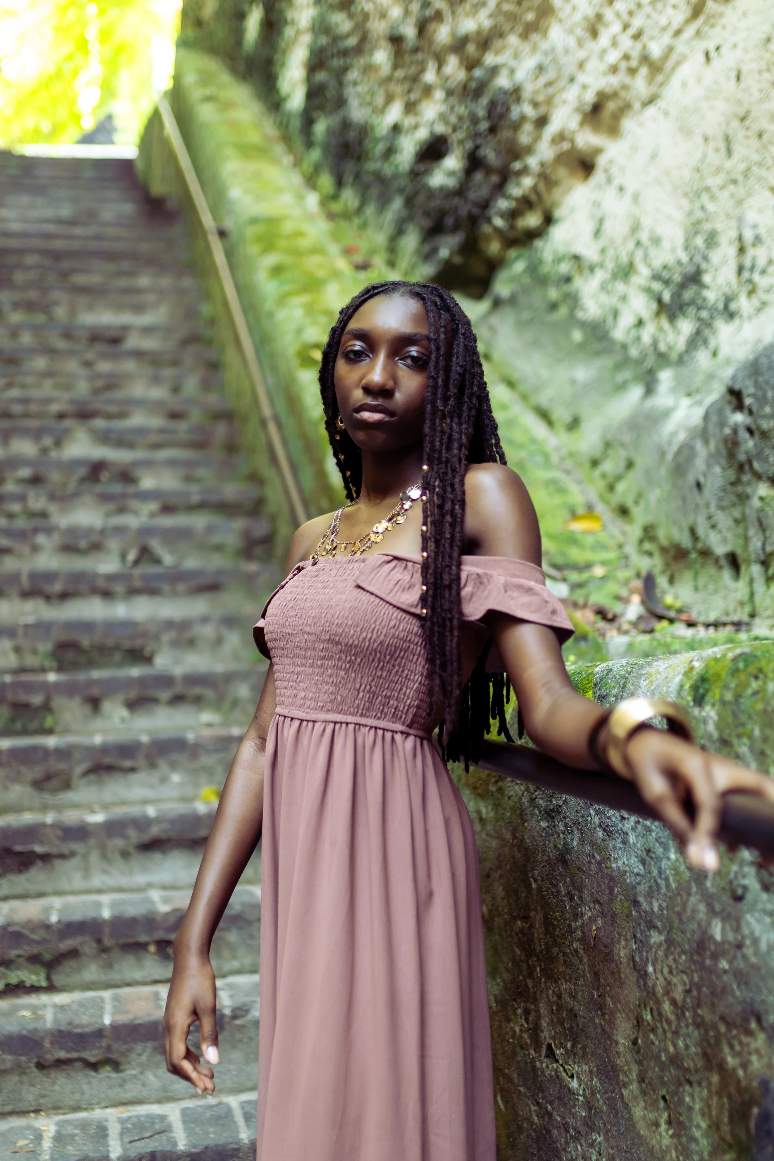 A woman with long braids, wearing a brown off-the-shoulder dress and jewelry, standing on outdoor stone stairs with a mossy rock wall and greenery in the background.