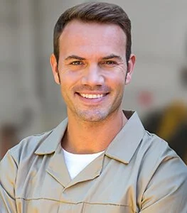 A man with short dark hair and a friendly smile, wearing a light-colored uniform with a white undershirt, standing outdoors.